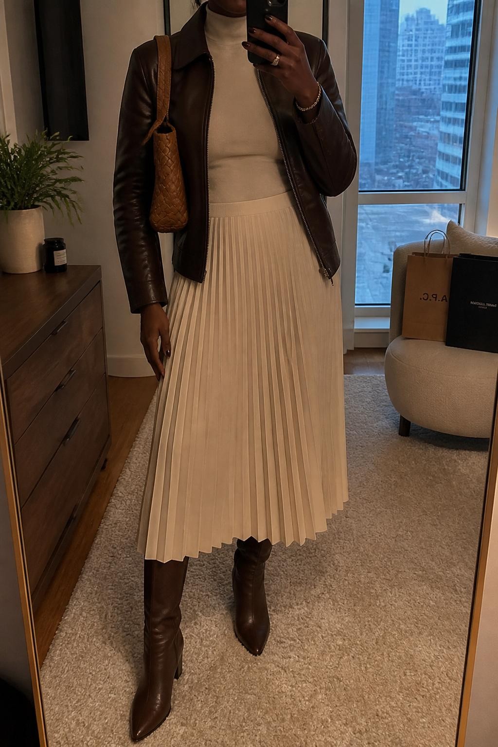 mirror selfie of a Black woman wearing a chocolate brown leather jacket, fitted ivory mock-neck top, cream pleated midi skirt, brown knee-high leather boots, and a woven tan shoulder bag, clearly showing the same clothing items, colors, fabrics, and accessories listed in the Outfit Pieces section, holding a phone in front of a mirror, cropped at NECK so the face does not show, with a relaxed casual selfie pose, in a stylish city apartment, warm moody lighting, including a small plant on a dresser and shopping bags beside a chair, latest iPhone photo quality.