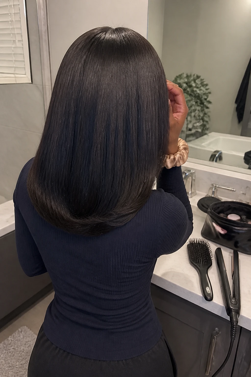 Image prompt

Photo of a woman wearing a medium-length straight weave with smooth strands and softly curved-under ends, back-of-head view highlighting the rounded shape, standing near a bathroom vanity mirror with a flat iron and paddle brush on the counter, soft indoor lighting, containing a small lived-in detail such as an open makeup bag and a silk scrunchie around her wrist, realistic getting-ready moment, latest iPhone photo quality.