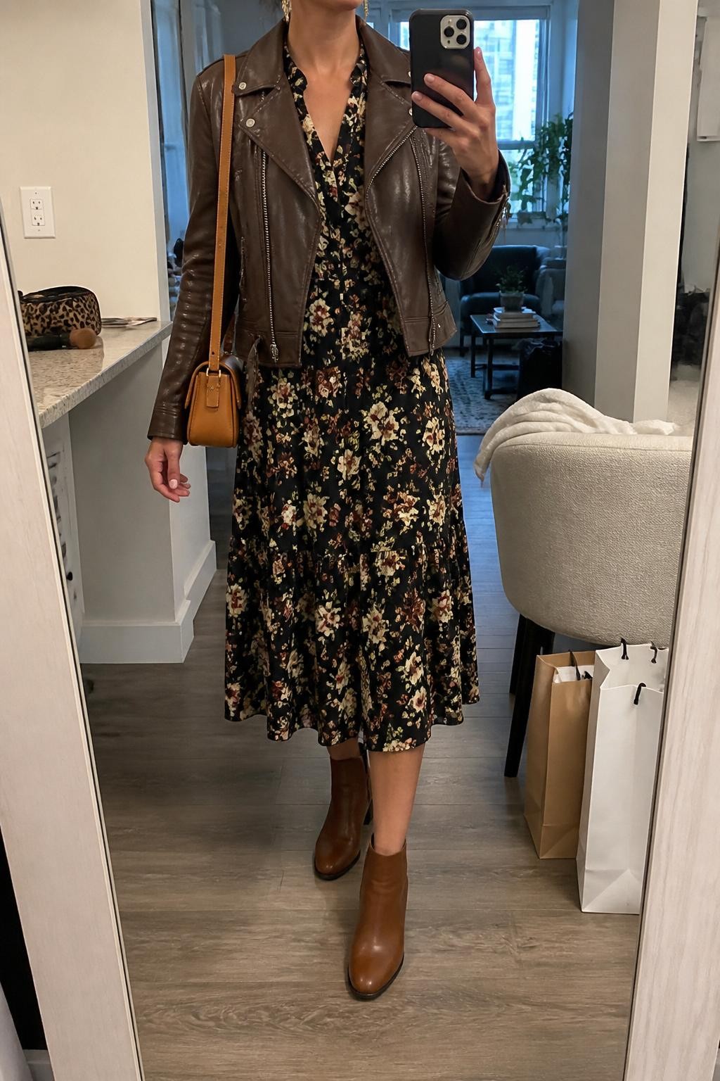 mirror selfie of a woman wearing the exact outfit described above, clearly showing a brown leather moto jacket layered over a black floral midi dress with cream and rust flowers, brown ankle boots, a tan mini shoulder bag, and gold drop earrings, holding a phone in front of a mirror, cropped at NECK so the face does not show, with a relaxed casual selfie pose, in a stylish city apartment, natural window light, with shopping bags beside a chair and a makeup brush or cosmetic bag on the counter, latest iPhone photo quality.