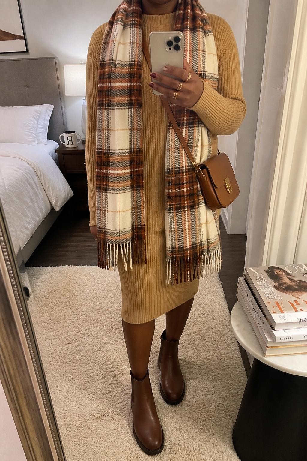 mirror selfie of a Black woman wearing the exact outfit described above, clearly showing a camel ribbed sweater dress, oversized plaid blanket scarf in cream rust and brown, brown Chelsea boots, tan crossbody bag, and gold stacked rings, holding a phone in front of a mirror, cropped at NECK so the face does not show, with a relaxed casual selfie pose, in a cozy bedroom mirror setup, soft indoor lighting, with a coffee mug on a bedside table and a stack of fashion magazines on a side table, latest iPhone photo quality.