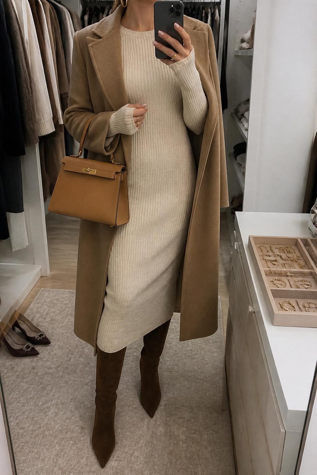 mirror selfie of a woman wearing the exact outfit described above, clearly showing chocolate brown suede knee-high boots, a cream ribbed sweater midi dress with long sleeves, a camel wool coat worn open, a tan structured handbag, and gold hoop earrings, holding a phone in front of a mirror, cropped at NECK so the face does not show, with a relaxed casual selfie pose, in a walk-in closet mirror, soft indoor lighting, with a jewelry tray on the vanity and a pair of heels on the floor nearby, latest iPhone photo quality.