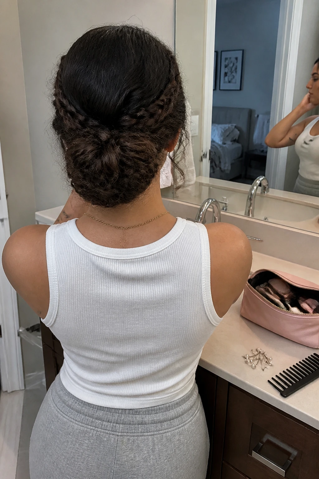 Image prompt

Photo of a woman wearing a braided curly bun updo with two textured side braids wrapped around a full low bun, back-of-head view highlighting the braid and bun details, standing at a bathroom vanity mirror, soft indoor lighting, containing a small lived-in detail such as pearl pins beside an open makeup bag and a wide-tooth comb on the counter, realistic getting-ready moment, latest iPhone photo quality.