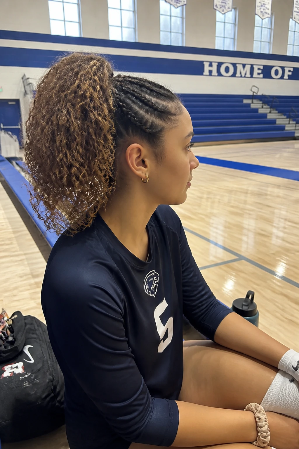 Image prompt

Photo of a woman wearing a high curly volleyball ponytail with two neat front braids feeding into the base, defined curls secured away from her face, candid side profile view inside a bright indoor volleyball gym, natural daylight mixed with soft court lighting, containing a small lived-in detail such as a sports bag on the bleachers, a spare scrunchie on her wrist, and a water bottle near the court, realistic pre-game getting-ready moment, latest iPhone photo quality.