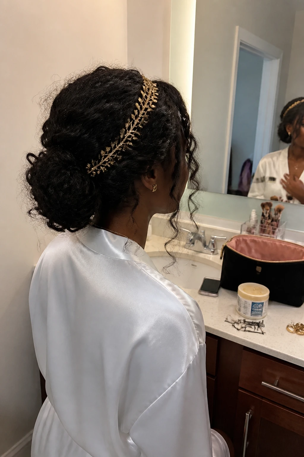 Image prompt

Photo of a Black woman wearing a soft low curly chignon with gentle crown volume, loose face-framing curls, and a delicate gold leaf headband, back three-quarter view beside a bathroom vanity mirror, soft indoor lighting, containing a small lived-in detail such as bobby pins beside an open makeup bag, a curl cream jar on the counter, and small gold earrings nearby, realistic formal getting-ready moment, latest iPhone photo quality.