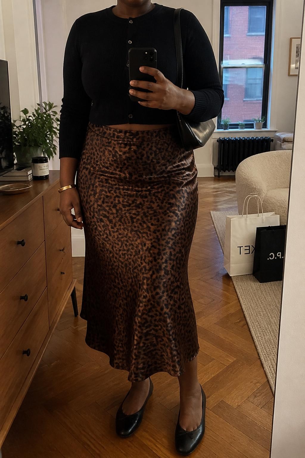 mirror selfie of a plus size Black woman wearing the exact outfit described above, a black soft knit cropped cardigan, brown satin leopard midi skirt with subtle sheen, black leather ballet flats, glossy black shoulder bag, and gold bangle bracelet, clearly showing the same clothing items, colors, fabrics, and accessories listed in the Outfit Pieces section, holding a phone in front of a mirror, cropped at NECK so the face does not show, with a relaxed casual selfie pose, in a stylish city apartment, warm moody lighting, including a small plant on a dresser and shopping bags beside a chair, latest iPhone photo quality.
