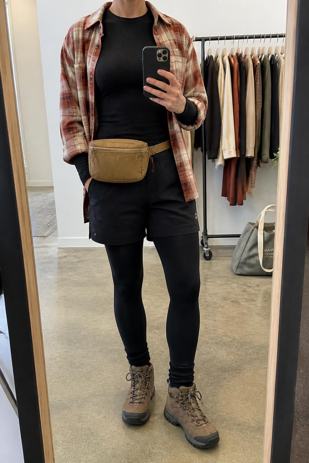mirror selfie of a woman wearing the exact outfit described above, clearly showing a rust and cream flannel shirt worn open, black fitted long-sleeve top, black hiking shorts, warm black fleece-lined tights, brown lace-up hiking boots, and a tan belt bag worn at the waist, holding a phone in front of a mirror, cropped at NECK so the face does not show, with a relaxed casual selfie pose, in a fashion studio mirror corner, soft indoor lighting, with a clothing rack with garments behind and a tote bag resting on the floor, latest iPhone photo quality.