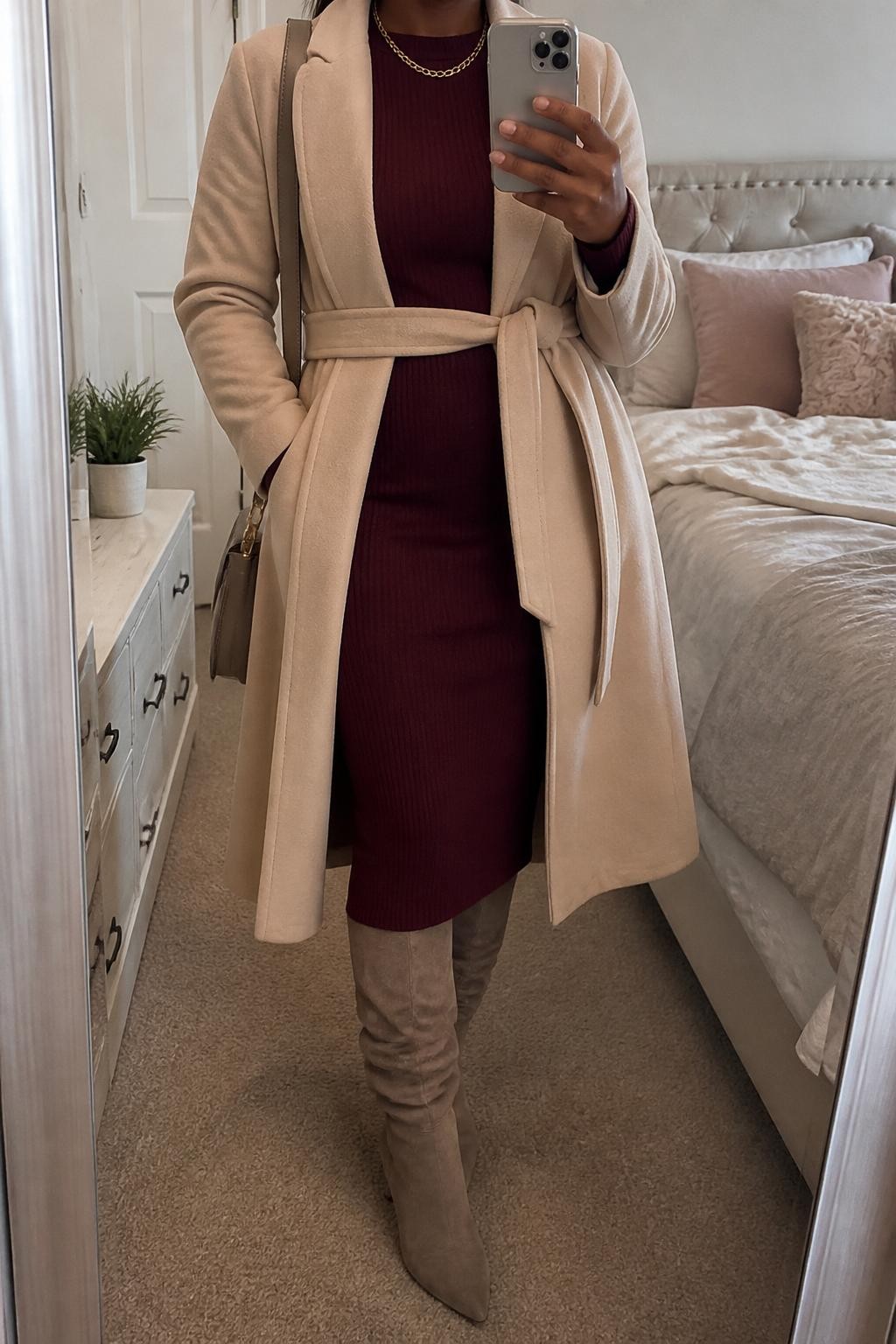 mirror selfie of a Black woman wearing the exact outfit described above, clearly showing taupe slouchy knee-high boots, a burgundy long-sleeve knit midi dress, a beige belted coat, a taupe shoulder bag, and a gold chain necklace, holding a phone in front of a mirror, cropped at NECK so the face does not show, with a relaxed casual selfie pose, in a feminine-themed bedroom, soft indoor lighting, with soft pillows on an unmade bed and a small plant on a dresser, latest iPhone photo quality.