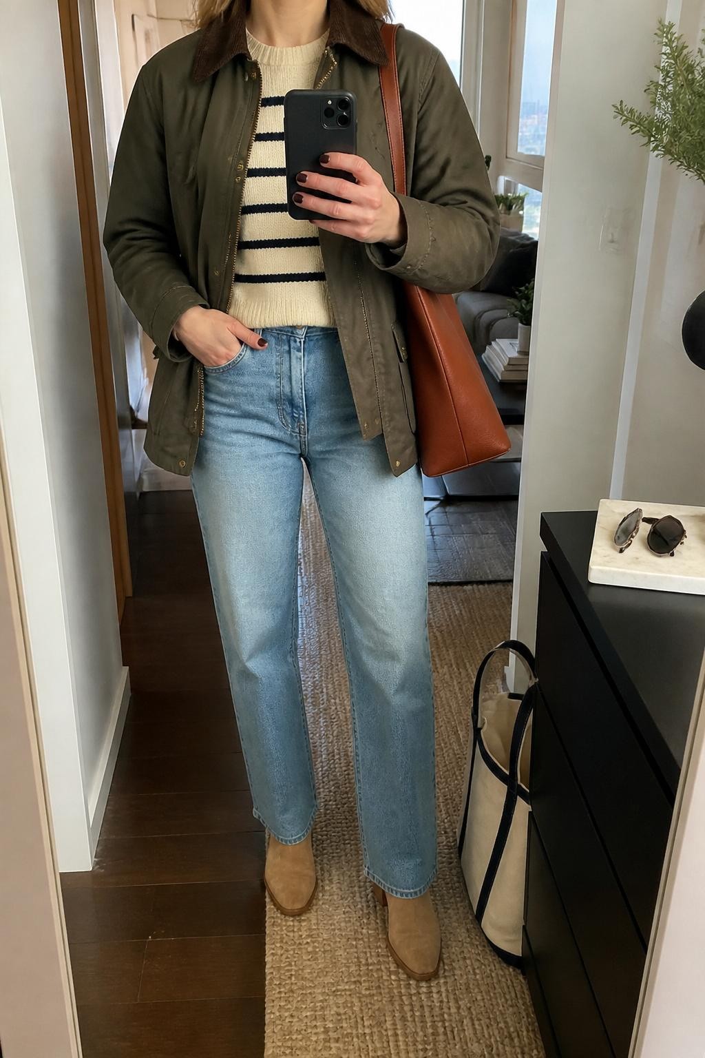 mirror selfie of a woman wearing the exact outfit described above, clearly showing an olive barn jacket, cream and navy striped knit sweater, light-wash straight-leg jeans, tan suede ankle boots, and a brown leather tote bag, holding a phone in front of a mirror, cropped at NECK so the face does not show, with a relaxed casual selfie pose, in a stylish city apartment, soft indoor lighting, with a tote bag resting on the floor and sunglasses placed on a dresser, latest iPhone photo quality.