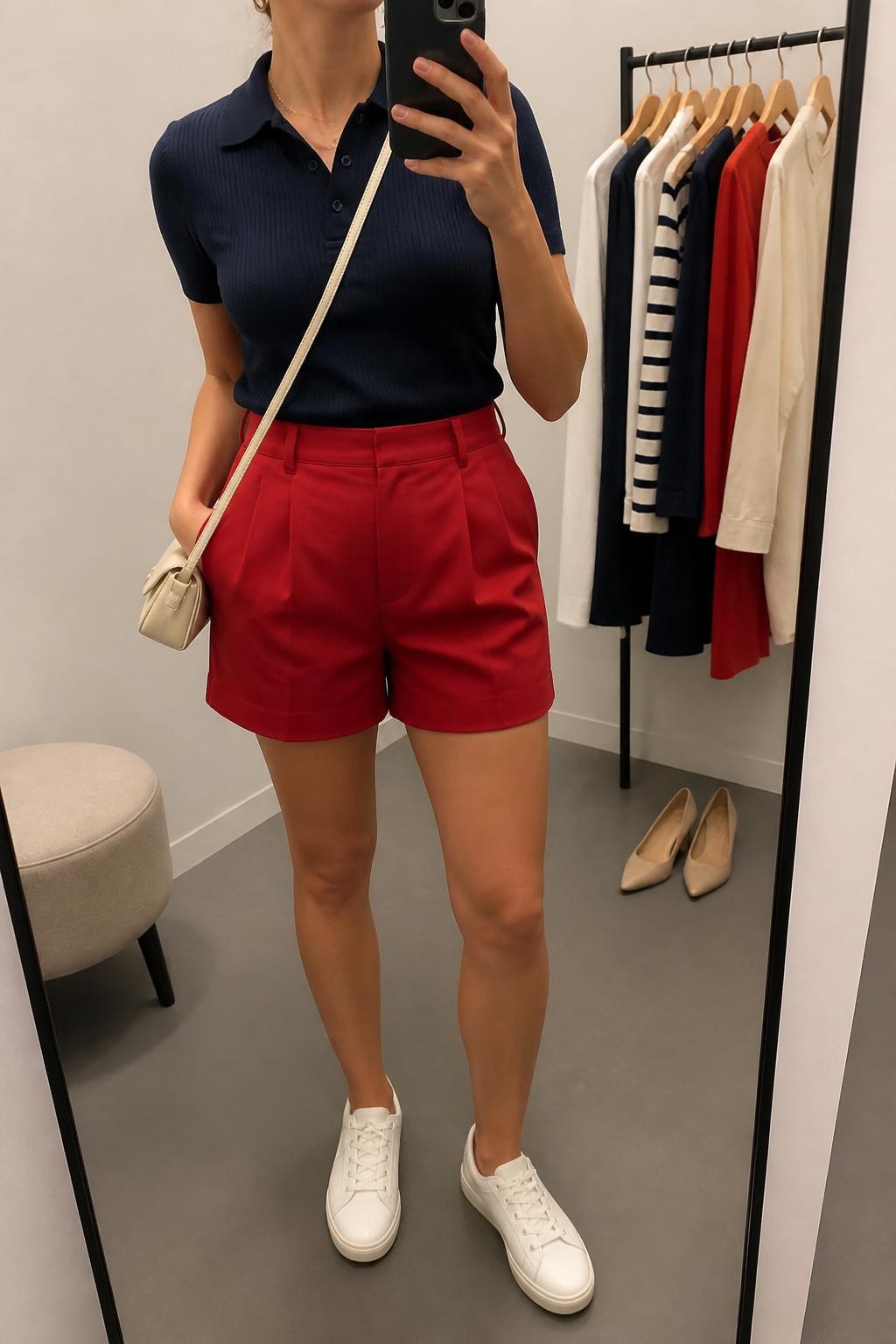 mirror selfie of a woman wearing a fitted navy short-sleeve knit polo shirt tucked into red high-waisted tailored shorts, white leather sneakers, cream mini crossbody bag, and small gold hoop earrings, clearly showing the same clothing items, colors, fabrics, and accessories listed in the Outfit Pieces section, holding a phone in front of a mirror, cropped at NECK so the face does not show, with a relaxed casual selfie pose, in a minimal dressing room, soft indoor lighting, including a clothing rack with garments behind and a pair of heels on the floor nearby, latest iPhone photo quality.