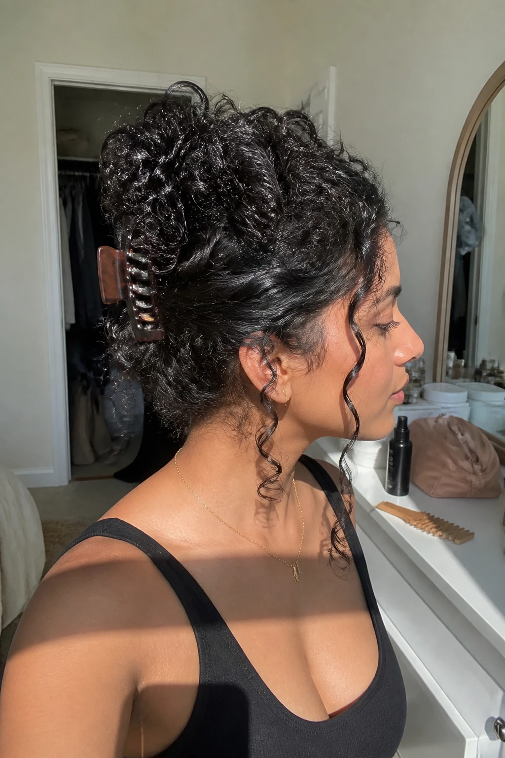 Image prompt

Photo of a South Asian woman wearing long 4A curls in a claw clip updo with the hair twisted upward, dense coil ends spilling softly over a large tortoiseshell clip, and two face-framing curls left loose, side profile view in a sun-drenched bedroom, natural window light, containing a small lived-in detail such as a wide-tooth comb and curl spray bottle on the dresser, a makeup bag nearby, and a slightly open closet behind her, realistic getting-ready moment, latest iPhone photo quality.