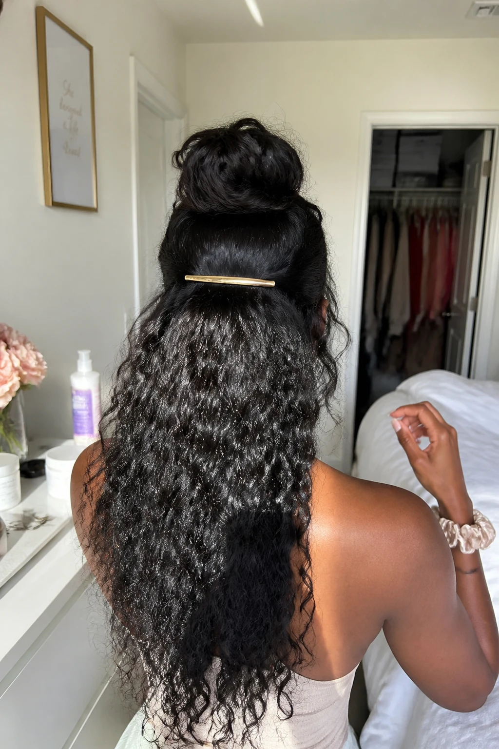 Image prompt
Photo of a Black woman wearing long curly sew-in hair in a half-up bun with a soft rounded shape at the crown, full loose curls cascading below, and a slim gold barrette placed beneath the bun, back-of-head view in a sun-drenched bedroom, natural window light, containing a small lived-in detail such as bobby pins and curl mousse on the dresser, a silk scrunchie on her wrist, and a slightly open closet behind her, realistic getting-ready moment, latest iPhone photo quality.