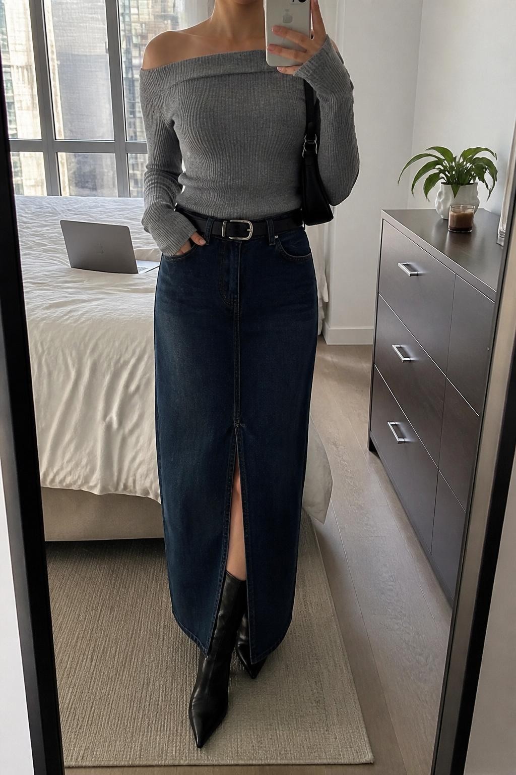 mirror selfie of a woman wearing a soft gray off-shoulder knit top, dark-wash denim maxi skirt with a front slit, black pointed kitten-heel boots, a silver buckle belt, and a black mini shoulder bag, clearly showing the same clothing items, colors, fabrics, and accessories listed in the Outfit Pieces section, holding a phone in front of a mirror, cropped at NECK so the face does not show, with a relaxed casual selfie pose, in a stylish city apartment, natural window light, including a small plant on a dresser and a laptop on the bed, latest iPhone photo quality.