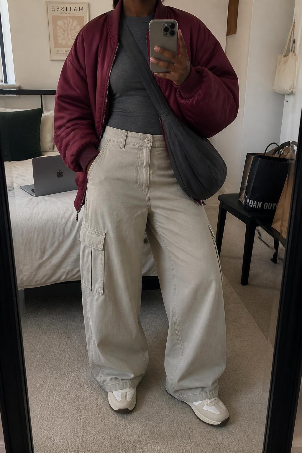Mirror selfie of a Black woman with deep brown skin wearing an oversized burgundy satin bomber jacket over a fitted charcoal gray long-sleeve top, high-waisted stone-colored wide-leg cargo trousers, cream retro-style sneakers, and a charcoal nylon crossbody bag, clearly showing the exact clothing items, colors, textures, and accessories listed, holding a latest-model iPhone in front of a mirror, cropped at the neck so the face does not show, relaxed casual selfie pose, in a cozy bedroom mirror setup with soft indoor lighting, a laptop on the bed and shopping bags beside a chair, natural uncluttered composition, latest iPhone photo quality.