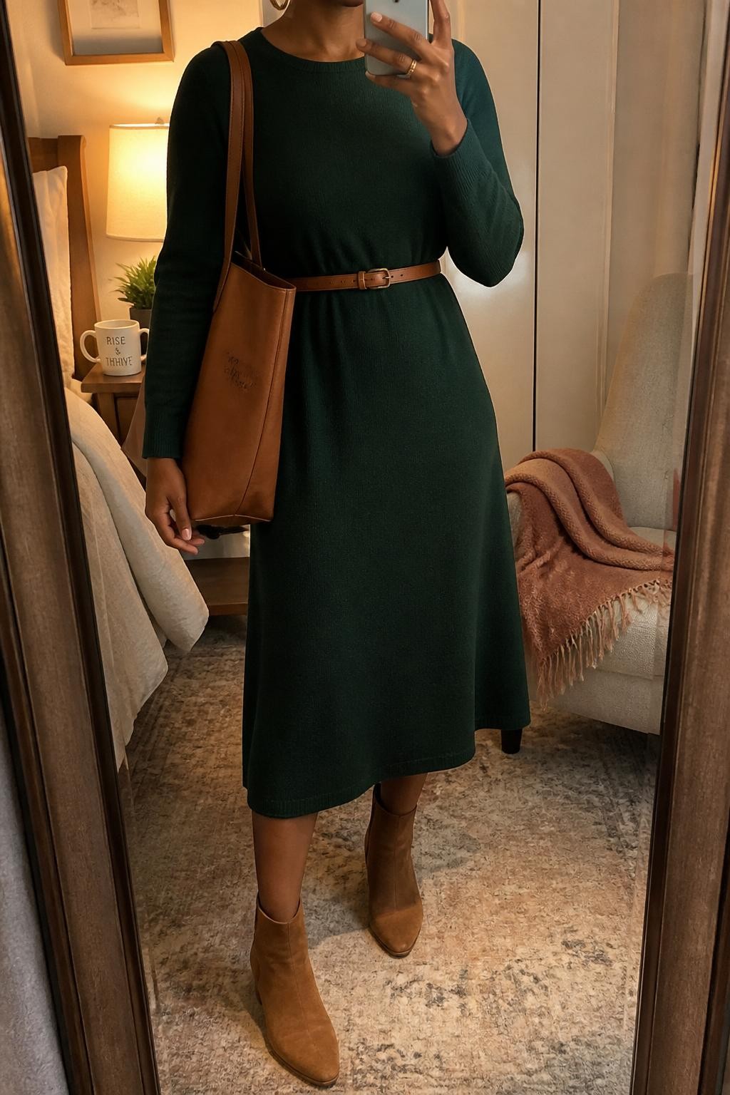 mirror selfie of a Black woman wearing a forest green long-sleeve midi dress, tan suede ankle boots, slim brown waist belt, gold hoop earrings, and a tan teacher tote, clearly showing the same clothing items, colors, fabrics, and accessories listed in the Outfit Pieces section, holding a phone in front of a mirror, cropped at NECK so the face does not show, with a relaxed casual selfie pose, in a cozy bedroom mirror setup, evening golden hour, including a folded blanket on a chair and a coffee mug on a bedside table, latest iPhone photo quality.