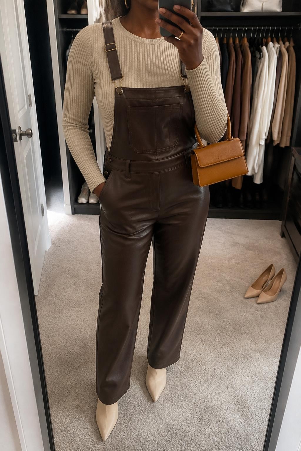 mirror selfie of a Black woman wearing dark chocolate faux leather overalls with a straight-leg cut over a fitted oatmeal ribbed knit top, cream pointed ankle boots, gold hoop earrings, and a structured tan mini bag, clearly showing the same clothing items, colors, fabrics, footwear, and accessories listed in the Outfit Pieces section, holding a phone in front of a mirror, cropped at NECK so the face does not show, with a relaxed casual selfie pose, in a walk-in closet mirror, soft indoor lighting, including an open closet with clothes visible and a pair of heels on the floor nearby, latest iPhone photo quality.