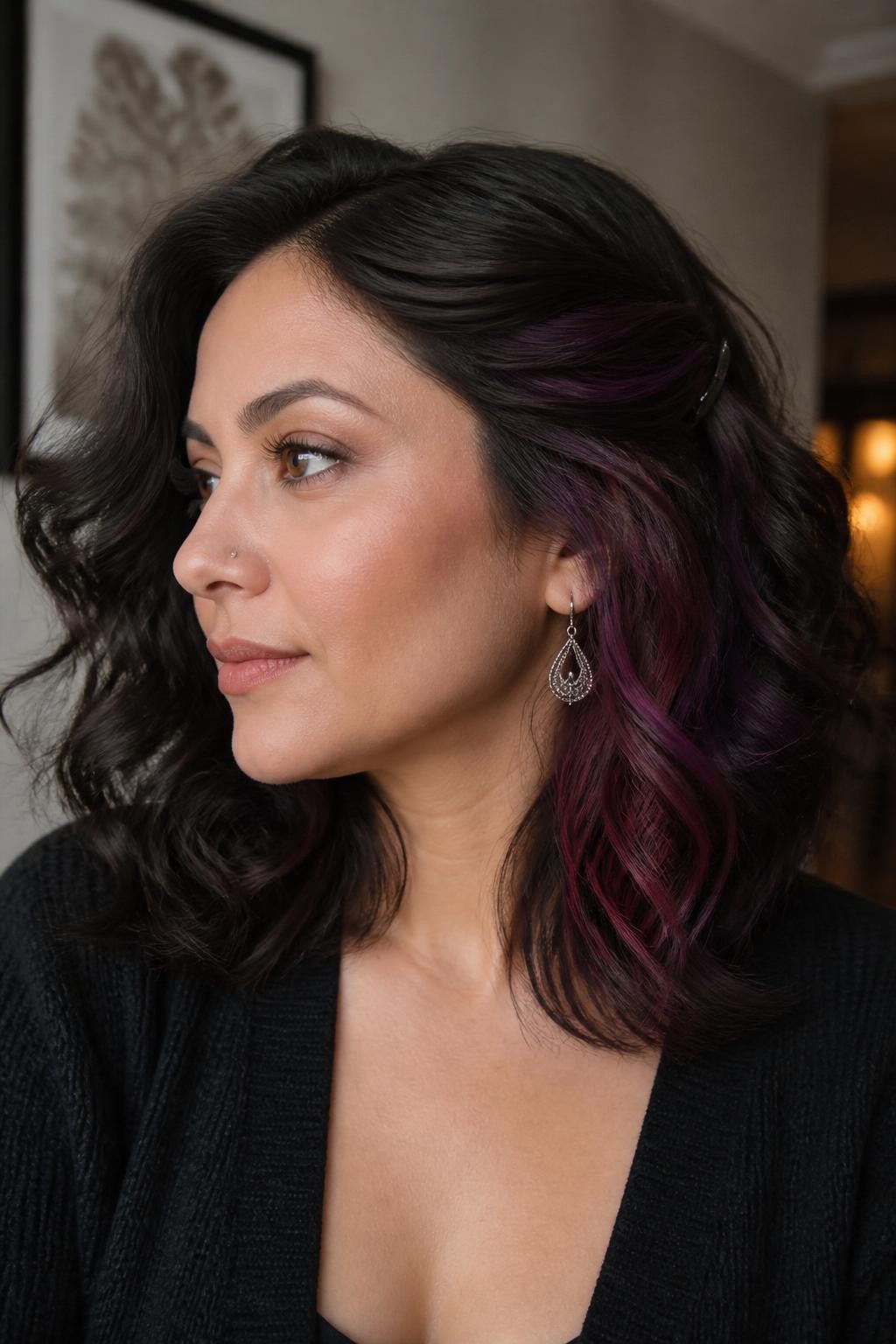 A realistic beauty portrait of a woman with medium skin and shoulder-length dark brunette curls, mulled plum underlights, neutral dark brown surface color, hidden panels of deep plum, blackberry, and wine-violet placed beneath both sides and throughout the lower nape, natural roots preserved inside the colored sections, one side pinned back to reveal the berry underlayer, upper curls remaining fully brunette, no bright purple or magenta blocks, textured black cardigan, silver earrings, warm indoor background, soft side lighting, realistic curl movement and precise hidden color placement.