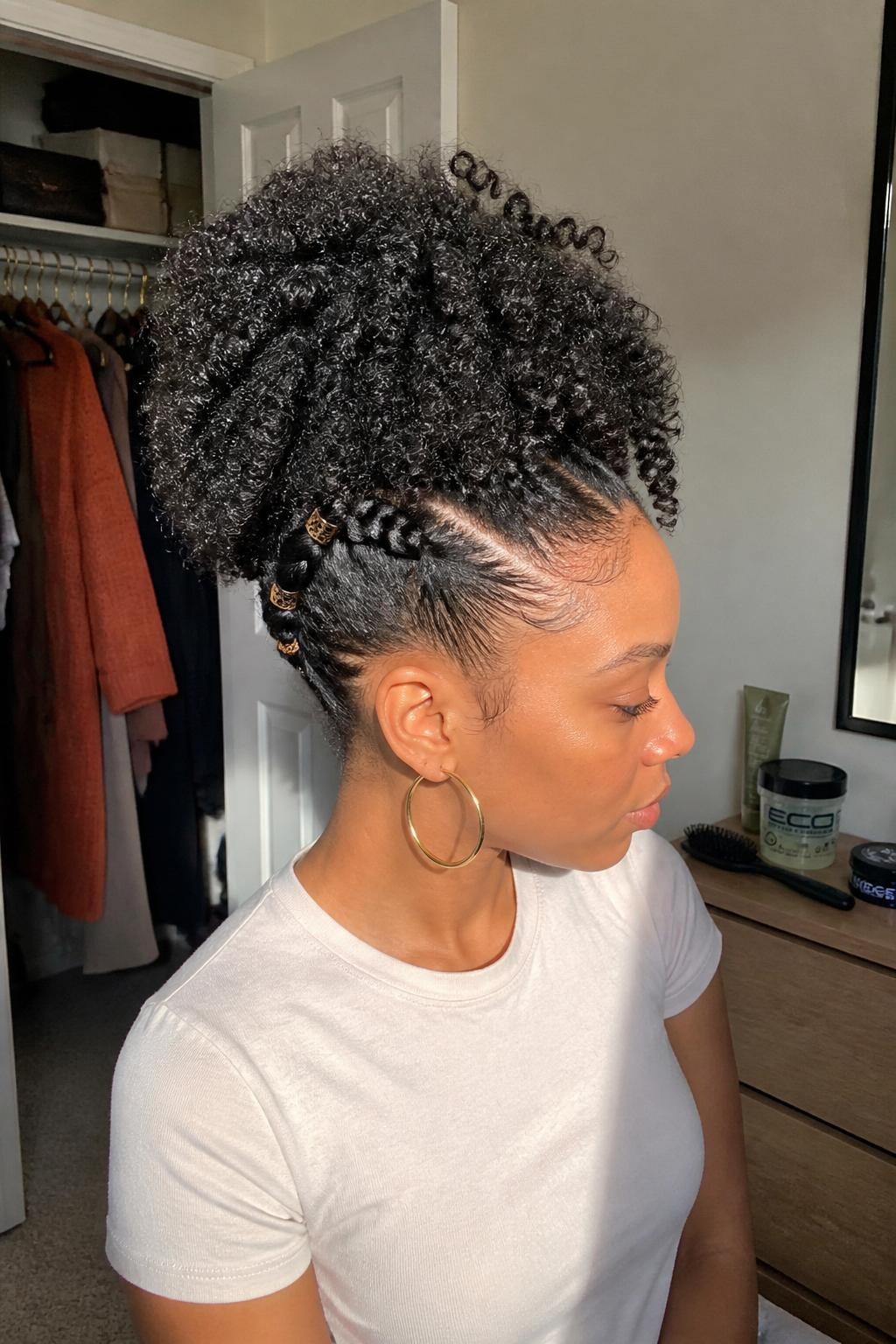 Photo of a Black woman wearing a full sculpted high puff with springy natural curls, two neat side twists leading toward the crown, softly shaped edges, and small bronze cuffs placed along the twists, side-profile view in a sun-drenched bedroom, warm natural window light, edge brush and curl gel on the dresser, rust cardigan hanging inside a slightly open closet and gold hoops peeking through, realistic natural texture, latest iPhone photo quality.