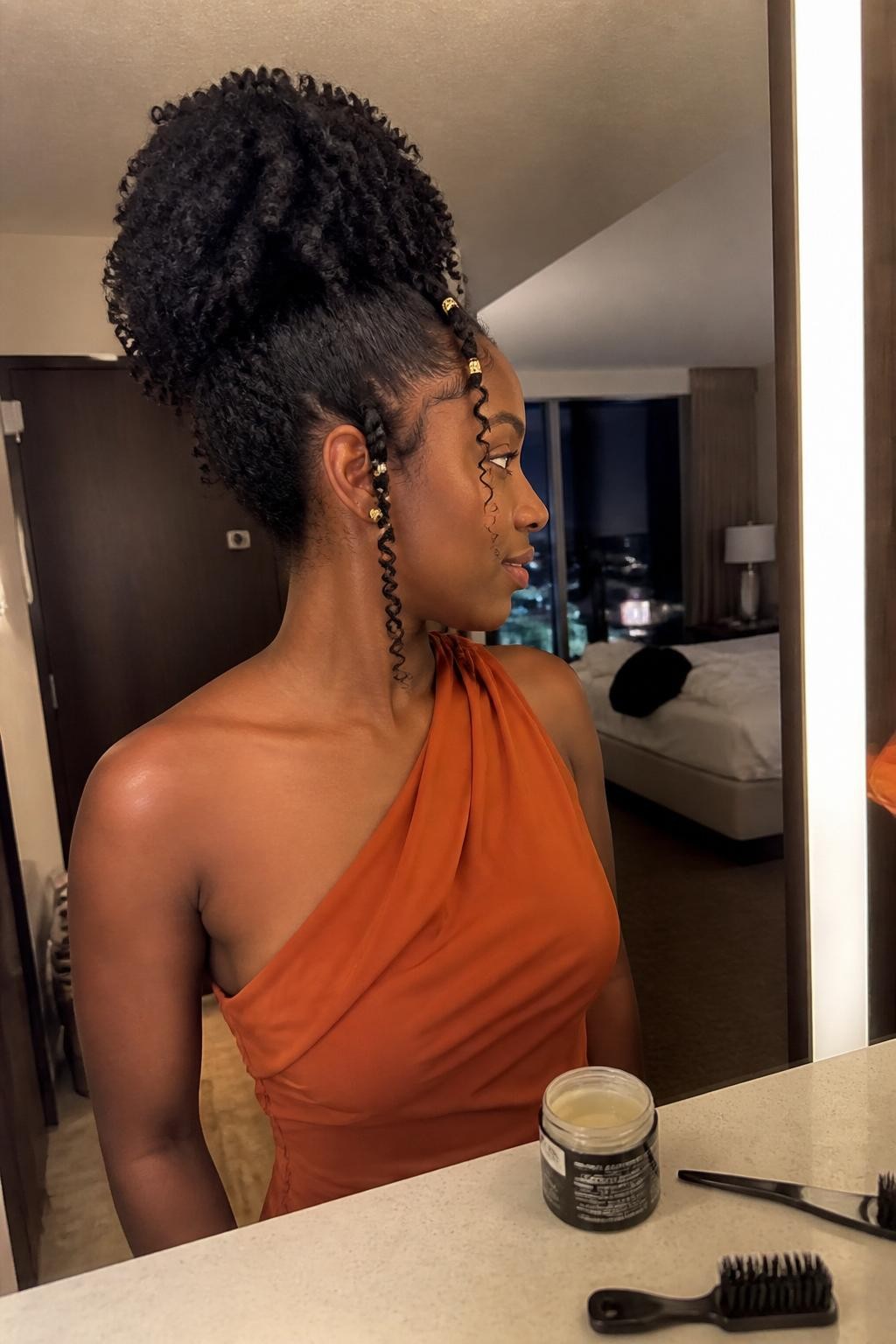 Photo of a Black woman wearing a full sculpted high puff with defined natural curls, gently laid edges, two curled tendrils near the temples, and small gold cuffs decorating slim side twists, candid side-profile mirror photo in a warm hotel suite, soft indoor lighting, edge brush and curl cream on the vanity, burnt-orange one-shoulder dress visible, realistic curl definition and natural texture, latest iPhone photo quality.