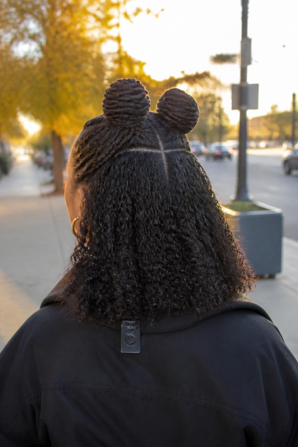 Image prompt: Photo of a Black woman wearing two small braided half-up space buns with defined shoulder-length curls left loose, back-of-head view showing the center part and braided bases, softly blurred city street background with fall foliage, golden hour glow, hair clip attached to her jacket collar and small hoop earrings peeking through, relaxed candid photo, realistic curl texture, latest iPhone photo quality.