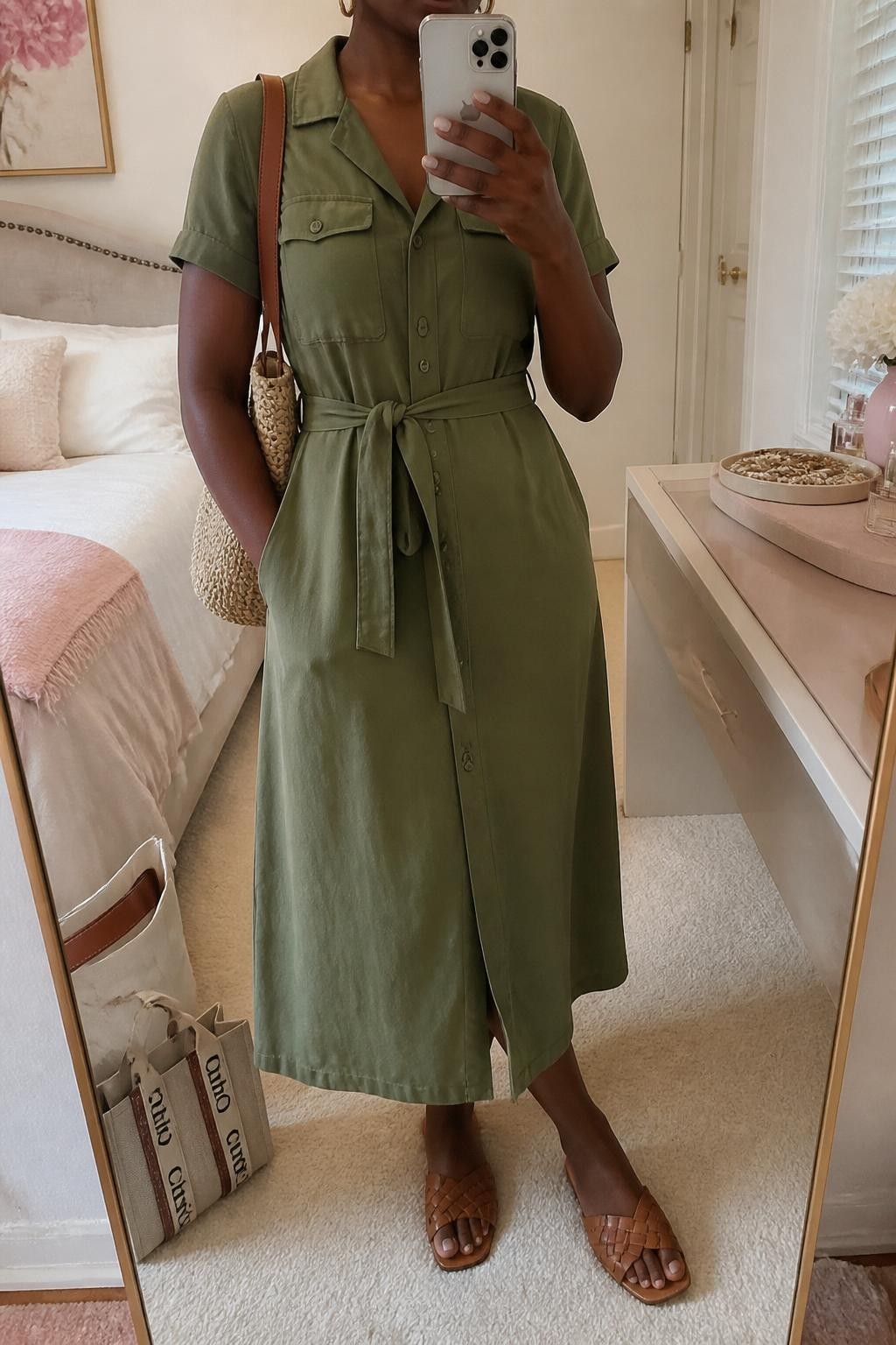 Mirror selfie of a Black woman with deep brown skin wearing an olive green lightweight cotton utility midi dress with short sleeves and a matching waist belt, flat woven tan leather sandals, a caramel raffia shoulder bag, and small gold hoop earrings, clearly showing the exact clothing items, colors, textures, and accessories listed, holding a latest-model iPhone in front of a mirror, cropped at the neck so the face does not show, relaxed casual selfie pose, in a feminine-themed bedroom with soft indoor lighting, a tote bag resting on the floor and a jewelry tray on the vanity, natural unstaged composition, latest iPhone photo quality.