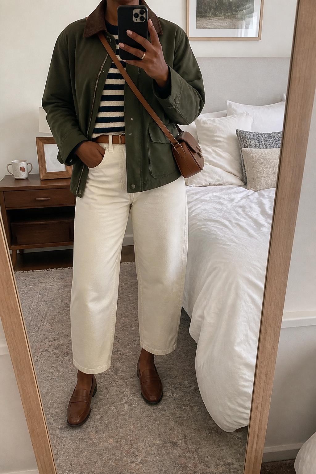 mirror selfie of a Black woman wearing the exact outfit described above, clearly showing an olive green barn jacket layered over a navy and cream striped sweater, cream barrel jeans, brown leather loafers, a cognac crossbody bag, and small gold stud earrings, holding a phone in front of a mirror, cropped at NECK so the face does not show, with a relaxed casual selfie pose, in a cozy bedroom mirror setup, soft indoor lighting, with a coffee mug on a bedside table and soft pillows on an unmade bed, latest iPhone photo quality.