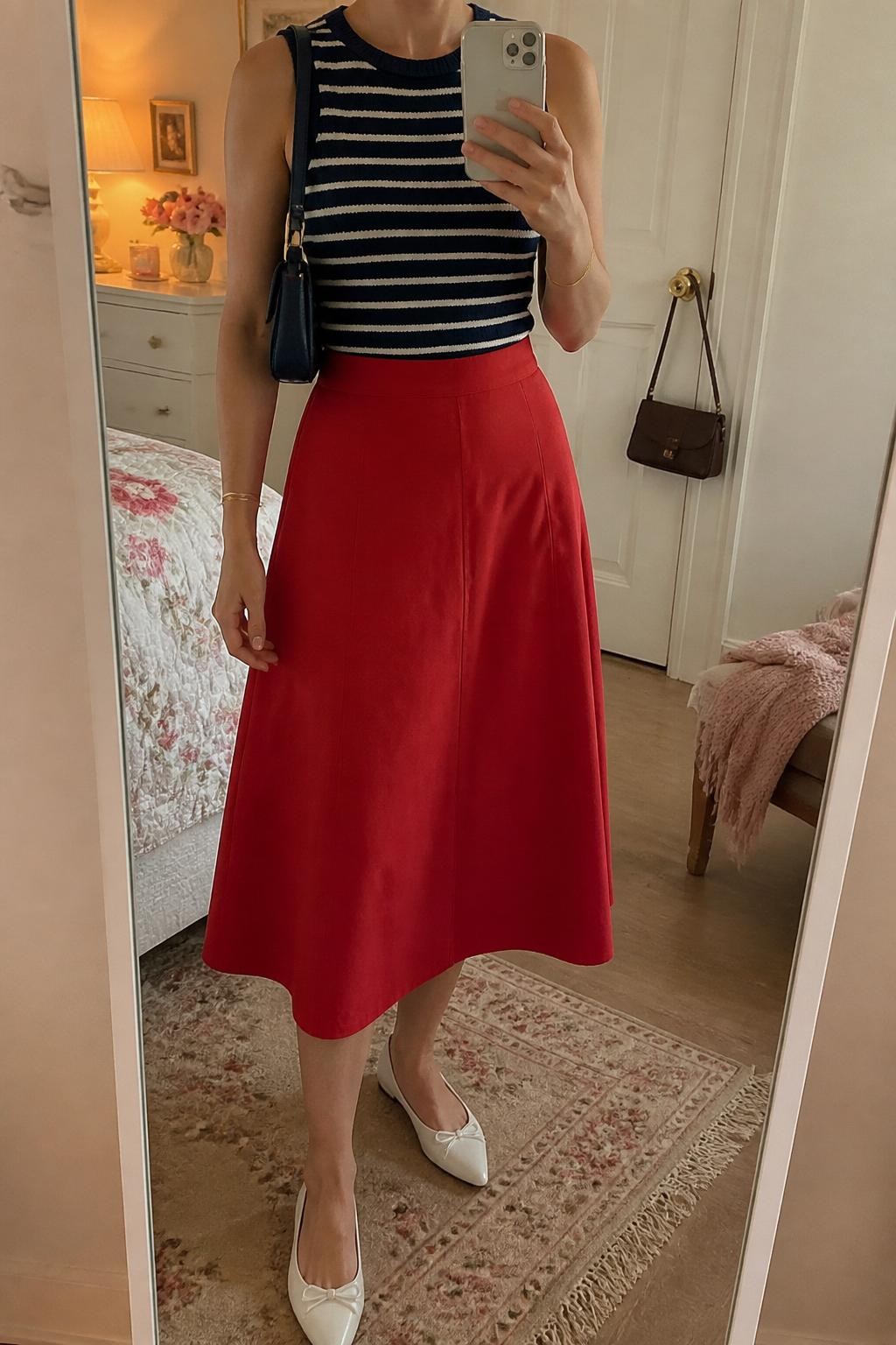 mirror selfie of a woman wearing a fitted navy and white striped sleeveless knit top tucked into a red cotton A-line midi skirt, white pointed ballet flats, navy mini shoulder bag, and gold bangle bracelet, clearly showing the same clothing items, colors, fabrics, and accessories listed in the Outfit Pieces section, holding a phone in front of a mirror, cropped at NECK so the face does not show, with a relaxed casual selfie pose, in a feminine-themed bedroom, evening golden hour, including a folded blanket on a chair and a handbag hanging on a door handle, latest iPhone photo quality.
