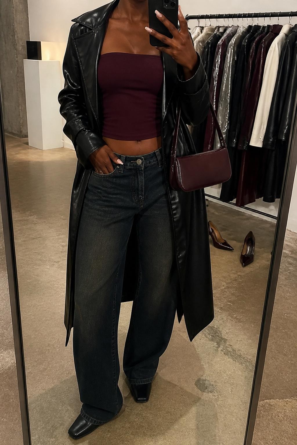mirror selfie of a Black woman wearing a burgundy fitted tube top, black faux leather trench coat, dark-wash low-rise wide-leg jeans, black square-toe boots, and a burgundy mini shoulder bag, clearly showing the same clothing items, colors, fabrics, and accessories listed in the Outfit Pieces section, holding a phone in front of a mirror, cropped at NECK so the face does not show, with a relaxed casual selfie pose, in a fashion studio mirror corner, warm moody lighting, including a clothing rack with garments behind and a pair of heels on the floor nearby, latest iPhone photo quality.
