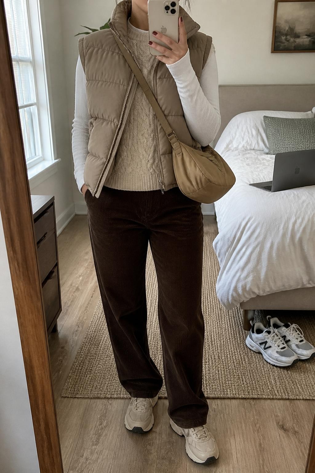 Mirror selfie of a woman wearing a quilted taupe puffer vest over an oatmeal cable-knit sweater and a fitted white thermal long-sleeve top, chocolate brown corduroy pants, cream hiking-inspired sneakers, and a tan nylon crossbody bag, clearly showing the exact clothing items, colors, textures, and accessories listed, holding a latest-model iPhone in front of a mirror, cropped at the neck so the face does not show, relaxed casual selfie pose, in a cozy bedroom mirror setup with natural window light, a laptop on the bed and a pair of sneakers on the floor nearby, natural uncluttered setting, latest iPhone photo quality.