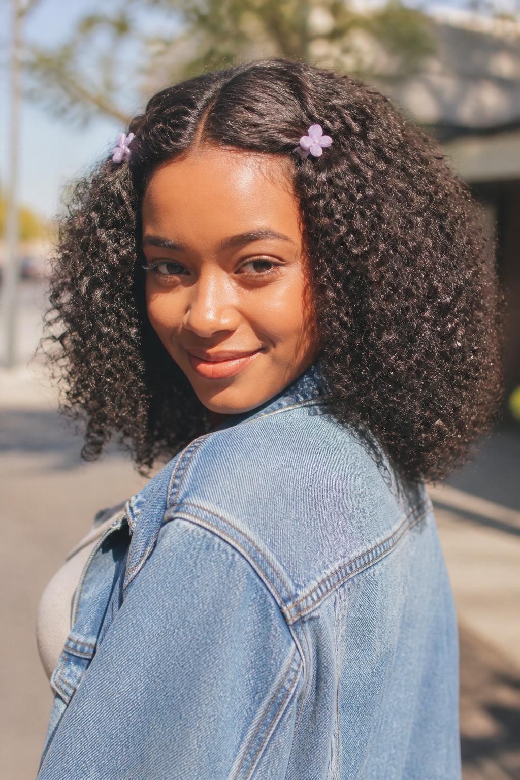 A realistic youthful hairstyle photo of a black woman with medium-length curly hair styled with two tiny pastel claw clips, one on each side near the temples, center part, defined curls left loose and rounded, warm brown skin, casual denim jacket, sunny outdoor street background, playful trendy curly hairstyle inspiration, bright natural light, high-resolution DSLR photo, no text, (no watermarks on images)