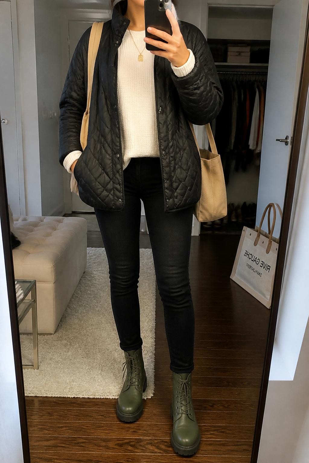mirror selfie of a woman wearing the exact outfit described above, clearly showing olive green combat boots, black skinny jeans, a cream thermal long-sleeve top, a black quilted jacket, a tan canvas tote bag, and a gold pendant necklace, holding a phone in front of a mirror, cropped at NECK so the face does not show, with a relaxed casual selfie pose, in a stylish city apartment, soft indoor lighting, with a tote bag resting on the floor and an open closet with clothes visible, latest iPhone photo quality.