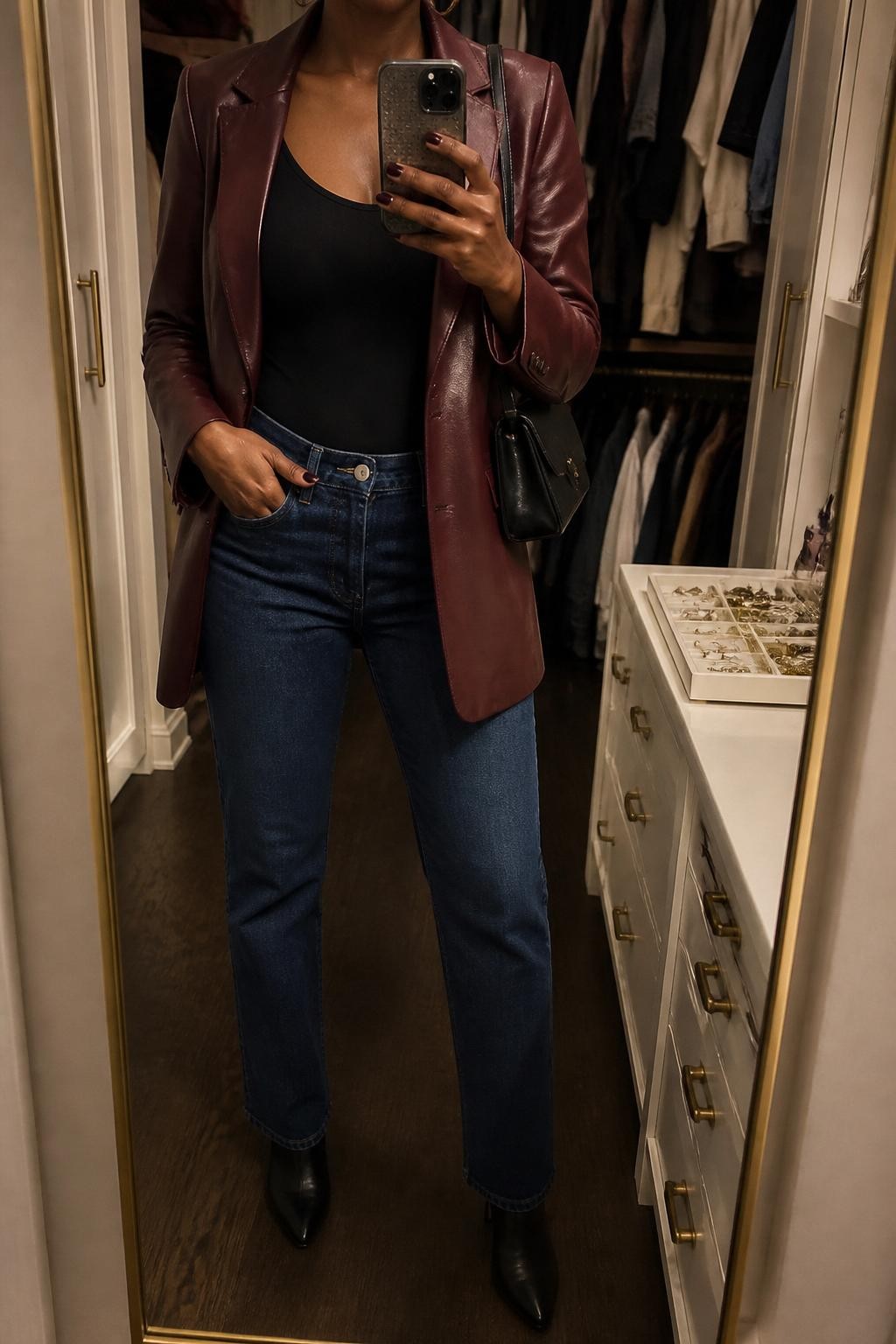 mirror selfie of a Black woman wearing the exact outfit described above, clearly showing a burgundy leather blazer layered over a black fitted bodysuit, dark blue straight-leg jeans, black heeled ankle boots, a black mini shoulder bag, and gold hoop earrings, holding a phone in front of a mirror, cropped at NECK so the face does not show, with a relaxed casual selfie pose, in a walk-in closet mirror, warm moody lighting, with an open closet with clothes visible and a jewelry tray on the vanity, latest iPhone photo quality.