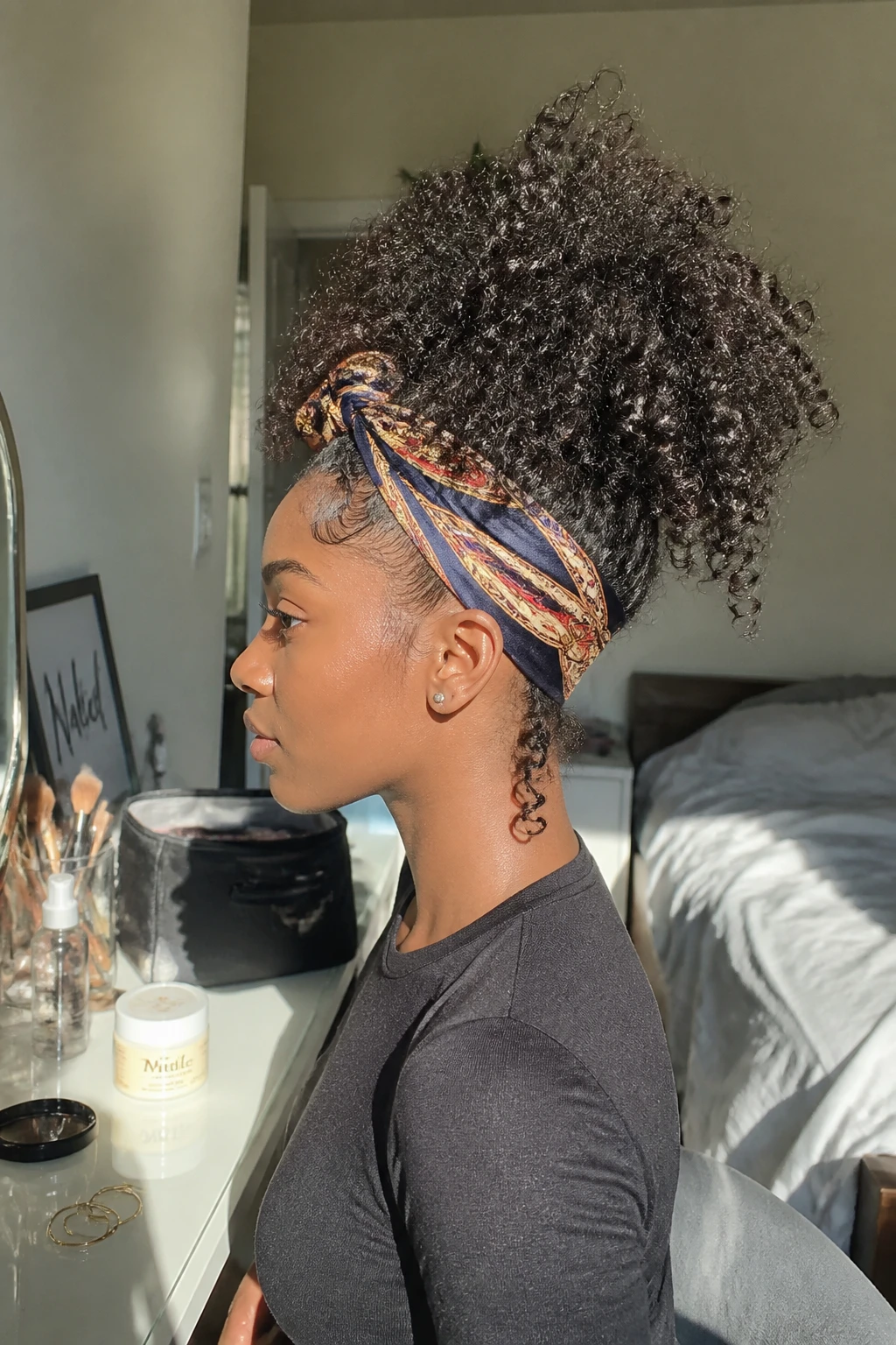 Image prompt
Photo of a Black woman wearing a high 3B curly pineapple with full springy ringlets gathered loosely at the crown and a narrow printed satin scarf tied around the hairline, side profile view inside a sun-drenched bedroom, natural window light, containing a small lived-in detail such as a curl cream jar on the vanity, hoop earrings nearby, and a makeup bag left open, realistic getting-ready moment, latest iPhone photo quality.