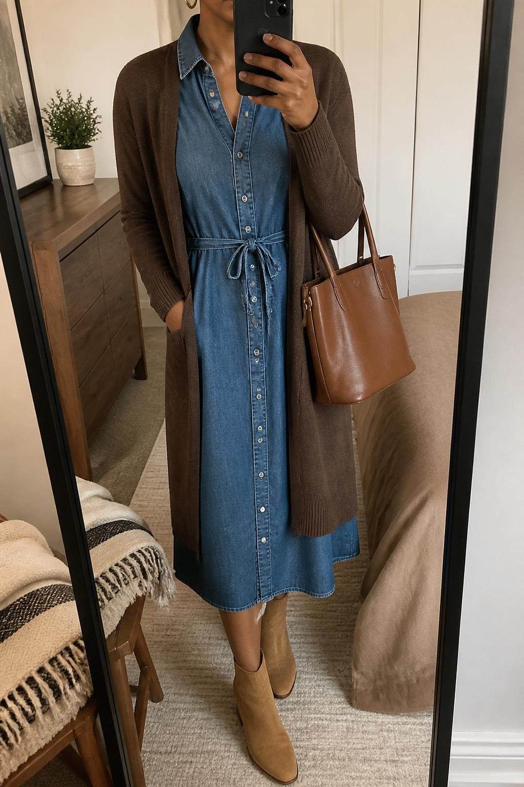 mirror selfie of a Black woman wearing a chambray button-front midi dress, chocolate brown long cardigan, tan ankle boots, small gold hoops, and a brown leather teacher bag, clearly showing the same clothing items, colors, fabrics, and accessories listed in the Outfit Pieces section, holding a phone in front of a mirror, cropped at NECK so the face does not show, with a relaxed casual selfie pose, in a cozy bedroom mirror setup, soft indoor lighting, including a folded blanket on a chair and a small plant on a dresser, latest iPhone photo quality.
