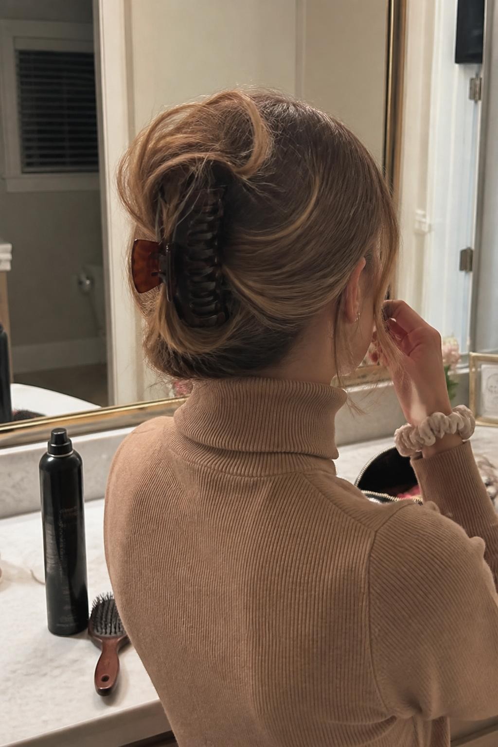 Photo of a woman wearing a collarbone-length lob folded into a relaxed French roll and held with a medium tortoiseshell claw clip, with softly curled ends spilling over the top and loose pieces around the ears, back-of-head view at a bathroom vanity, warm indoor lighting, texture spray and hairbrush beside an open makeup bag, camel turtleneck visible and a silk scrunchie around her wrist, realistic imperfect twist, latest iPhone photo quality.