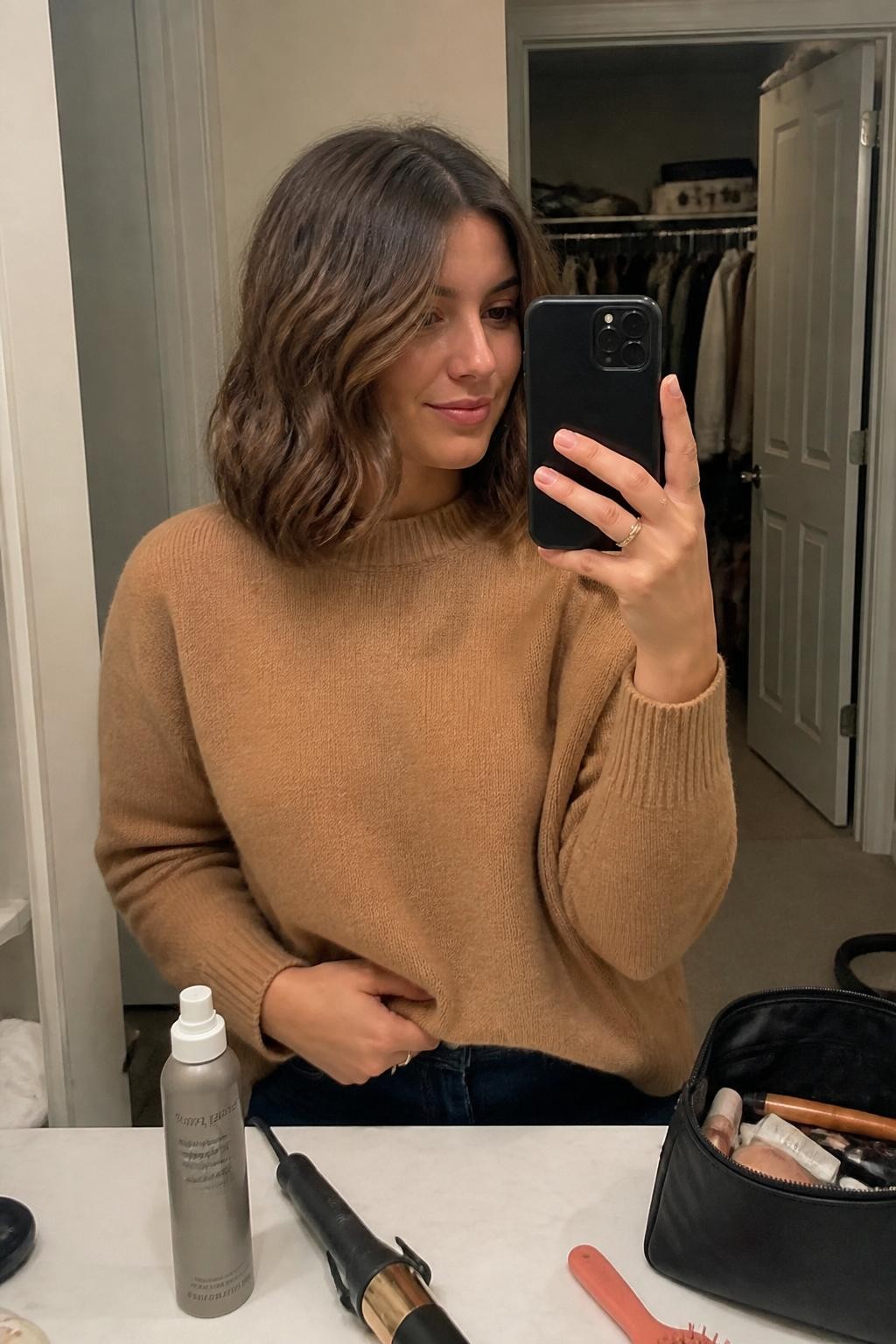 Photo of a woman wearing a collarbone-length chestnut-brown lob with loose ribbon-shaped waves, a soft center part, and lightly textured ends, candid mirror selfie at a bathroom vanity, warm indoor lighting, medium curling wand and heat-protectant spray resting beside an open makeup bag, camel knit sweater visible and a slightly open closet in the background, realistic flyaways and natural movement, latest iPhone photo quality.