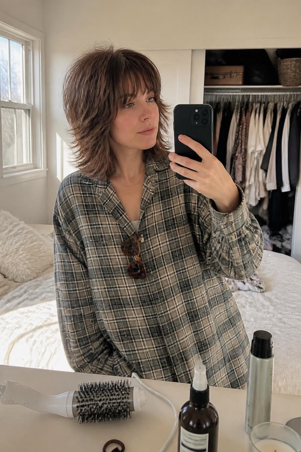 Photo of a woman wearing a shoulder-length shag lob with choppy crown layers, bottleneck bangs, and tousled outward-flicked ends, candid mirror selfie in a sun-drenched bedroom, natural window light, blow-dry brush and texture spray on the dresser, tortoiseshell clip attached to the collar of a plaid overshirt and a slightly open closet behind her, realistic separated layers, latest iPhone photo quality.