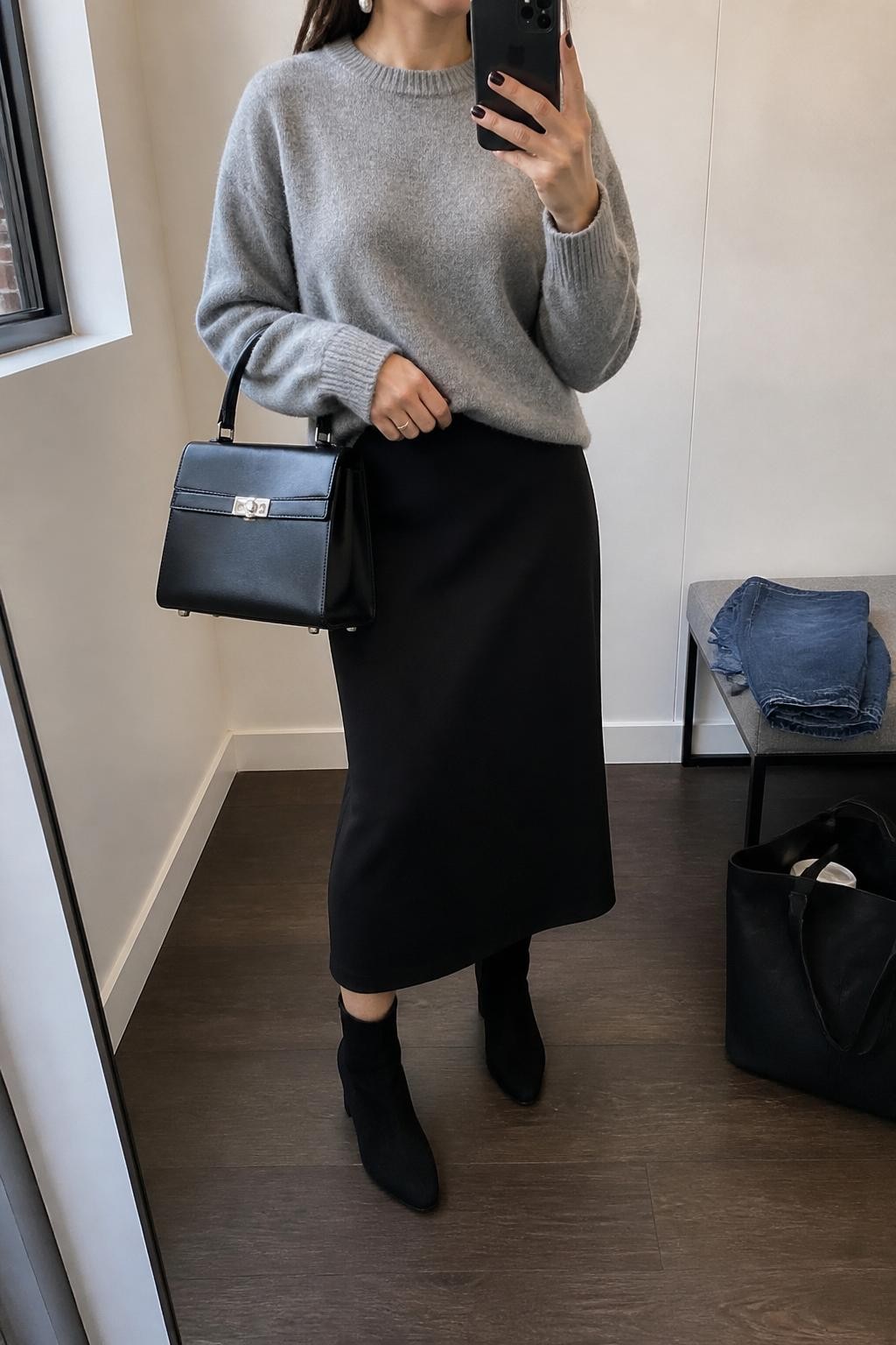 mirror selfie of a woman wearing a soft gray cashmere sweater, black wool midi skirt, black suede ankle boots, silver stud earrings, and a black leather top-handle bag, clearly showing the same clothing items, colors, fabrics, and accessories listed in the Outfit Pieces section, holding a phone in front of a mirror, cropped at NECK so the face does not show, with a relaxed casual selfie pose, in a minimal dressing room, natural window light, including a tote bag resting on the floor and folded denim jeans on a bench, latest iPhone photo quality.
