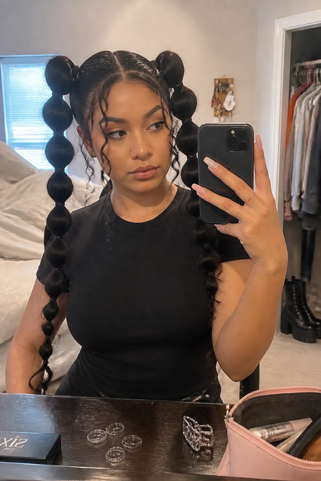 Image prompt
Photo of a Latina woman wearing two high curly bubble pigtails with a sharp zigzag middle part, compact rounded sections, and two loose spiral tendrils around the face, candid bedroom mirror selfie, natural window light, containing a small lived-in detail such as clear elastics on the dresser, black platform boots near the closet, and a silver claw clip beside a makeup bag, realistic alternative getting-ready moment, latest iPhone photo quality.