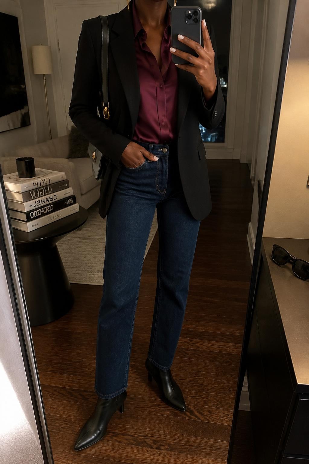 mirror selfie of a Black woman wearing the exact outfit described above, clearly showing black heeled ankle boots, dark blue straight-leg jeans, a burgundy satin blouse, a black tailored blazer, a black shoulder bag, and gold drop earrings, holding a phone in front of a mirror, cropped at NECK so the face does not show, with a relaxed casual selfie pose, in a stylish city apartment, warm moody lighting, with a stack of fashion magazines on a side table and sunglasses placed on a dresser, latest iPhone photo quality.