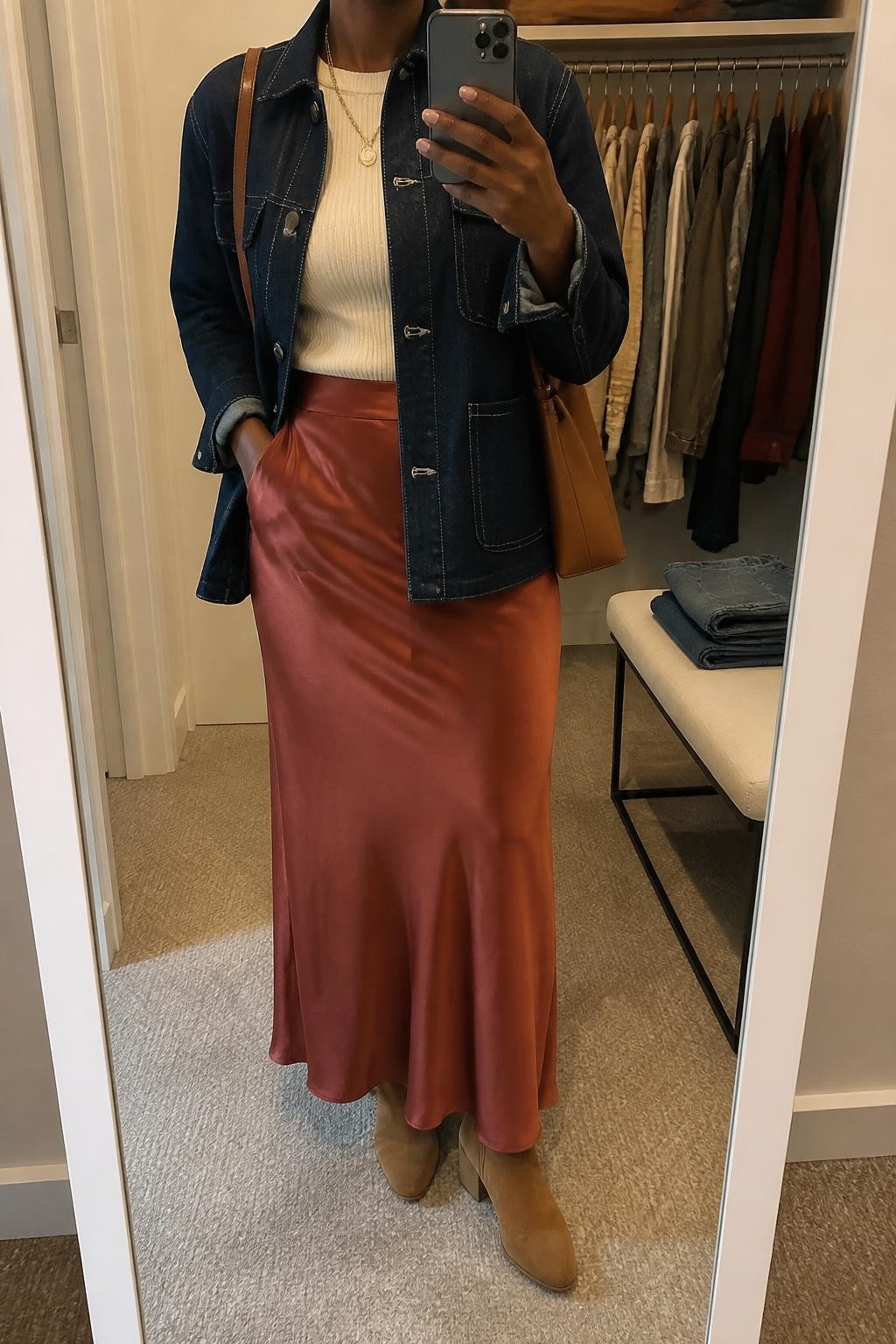 mirror selfie of a Black woman wearing the exact outfit described above, clearly showing a dark indigo denim chore jacket layered over a cream fitted knit top, rust satin maxi skirt, tan suede ankle boots, a brown leather shoulder bag, and gold layered necklaces, holding a phone in front of a mirror, cropped at NECK so the face does not show, with a relaxed casual selfie pose, in a walk-in closet mirror, soft indoor lighting, with an open closet with clothes visible and folded denim jeans on a bench, latest iPhone photo quality.