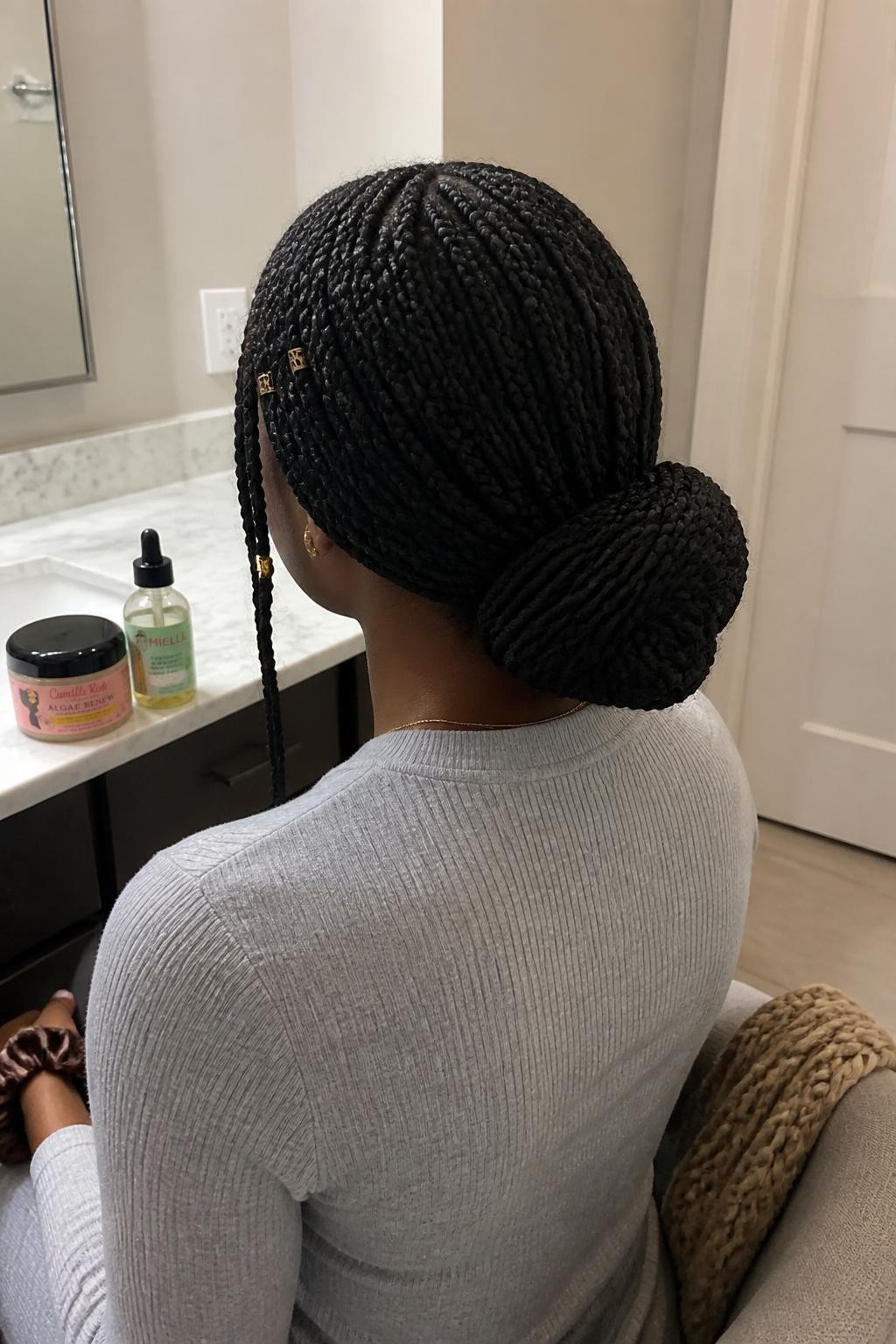 Photo of a Black woman wearing long knotless braids gathered into a wide low bun at the nape, with two slim curved braids left loose near the face and small gold cuffs placed near the temples, back-of-head view at a bathroom vanity, soft indoor lighting, braid mousse and scalp oil resting on the counter, a silk scrunchie around her wrist and a knit scarf draped over a nearby chair, realistic braided texture, latest iPhone photo quality.