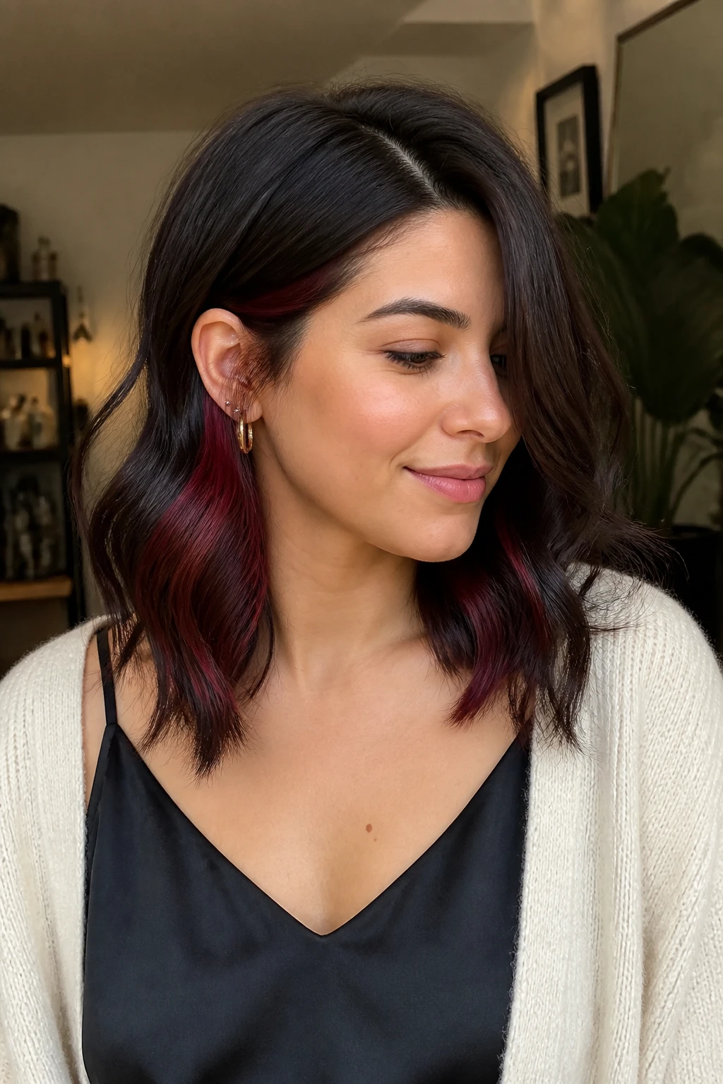 A realistic portrait of a woman with light-medium skin and dark brown shoulder-length hair, merlot brunette underlights, deep chocolate top layer, hidden wine-red pieces placed underneath near the nape and behind the ears, loose waves, one side tucked to reveal the merlot color, dark root shadow, black satin camisole under a soft cardigan, warm indoor light, realistic color placement, no bright fantasy red.