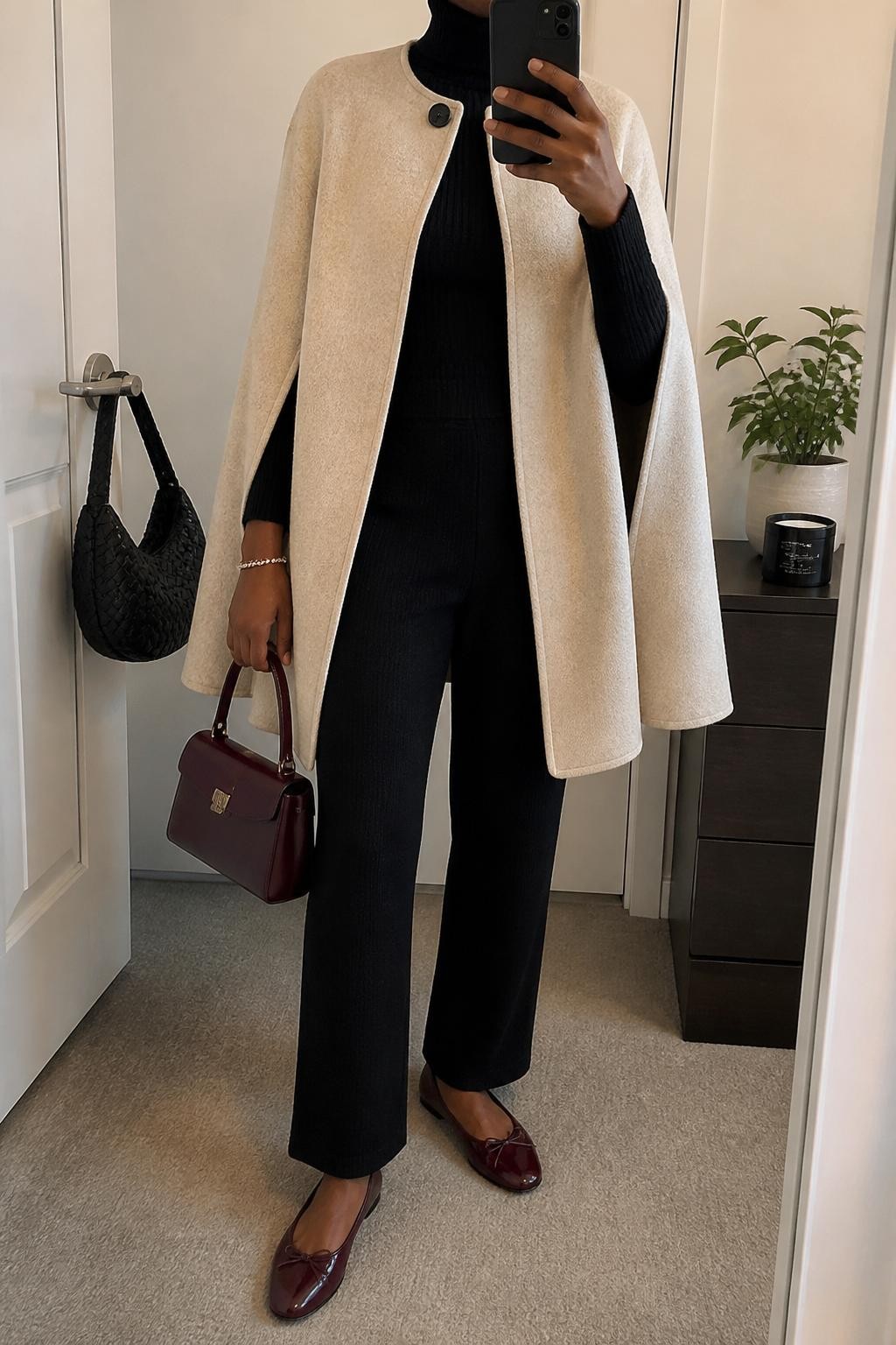 mirror selfie of a Black woman wearing an oatmeal wool cape coat, fitted black ribbed turtleneck, black straight-leg knit trousers, burgundy leather flats, and a burgundy structured mini bag, clearly showing the same clothing items, colors, fabrics, and accessories listed in the Outfit Pieces section, holding a phone in front of a mirror, cropped at NECK so the face does not show, with a relaxed casual selfie pose, in a minimal dressing room, soft indoor lighting, including a small plant on a dresser and a handbag hanging on a door handle, latest iPhone photo quality.