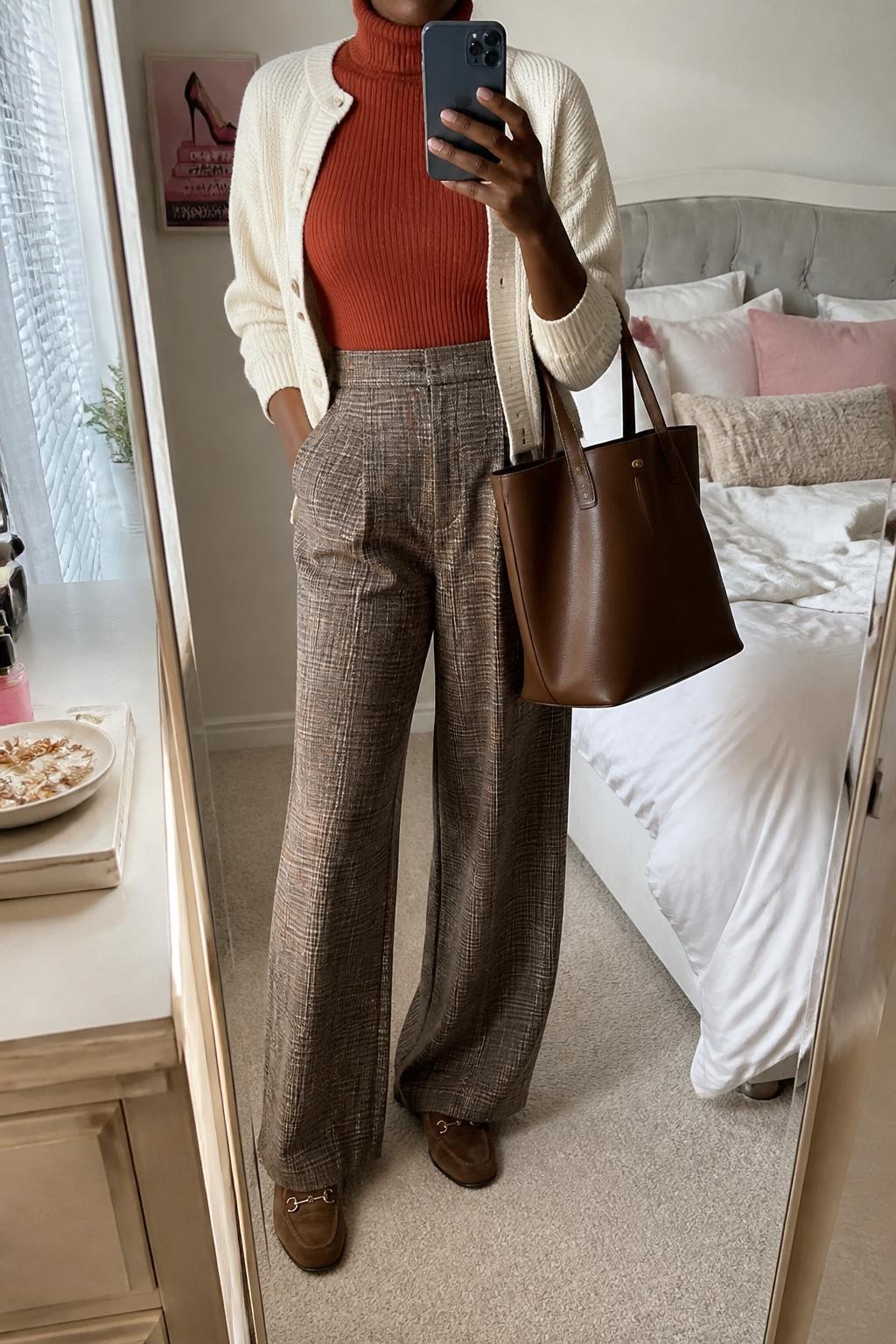 mirror selfie of a Black woman wearing a rust ribbed turtleneck, brown plaid wide-leg pants, cream cropped cardigan, brown suede loafers, and a structured brown work tote, clearly showing the same clothing items, colors, fabrics, and accessories listed in the Outfit Pieces section, holding a phone in front of a mirror, cropped at NECK so the face does not show, with a relaxed casual selfie pose, in a feminine-themed bedroom, natural window light, including soft pillows on an unmade bed and a jewelry tray on the vanity, latest iPhone photo quality.