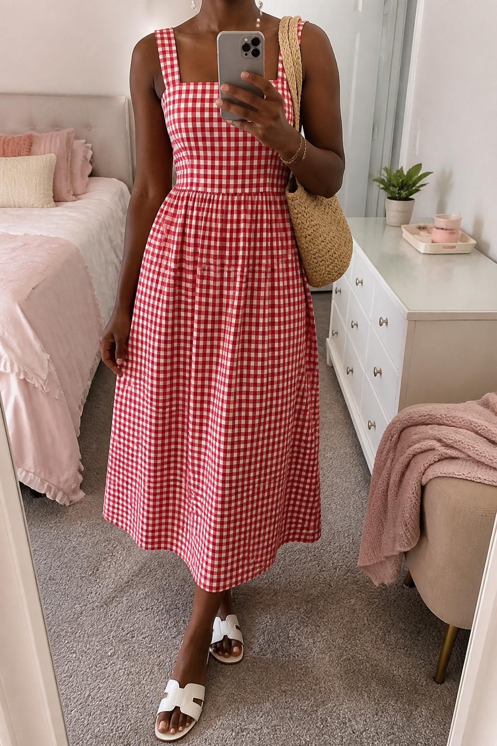 mirror selfie of a Black woman wearing a red and white gingham cotton midi dress with a fitted waist, white flat sandals, woven straw shoulder bag, pearl stud earrings, and a delicate gold bracelet, clearly showing the same clothing items, colors, fabrics, and accessories listed in the Outfit Pieces section, holding a phone in front of a mirror, cropped at NECK so the face does not show, with a relaxed casual selfie pose, in a feminine-themed bedroom, soft indoor lighting, including a small plant on a dresser and a folded blanket on a chair, latest iPhone photo quality.