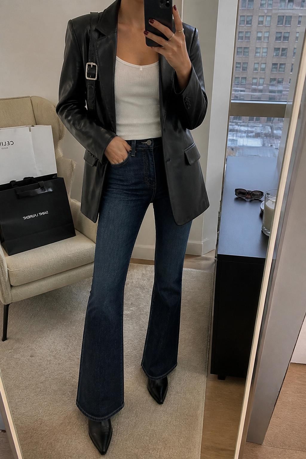 mirror selfie of a woman wearing a black leather blazer, fitted white ribbed tank top, dark-wash flare jeans, black pointed ankle boots, and a silver buckle shoulder bag, clearly showing the same clothing items, colors, fabrics, and accessories listed in the Outfit Pieces section, holding a phone in front of a mirror, cropped at NECK so the face does not show, with a relaxed casual selfie pose, in a stylish city apartment, soft indoor lighting, including shopping bags beside a chair and sunglasses placed on a dresser, latest iPhone photo quality.