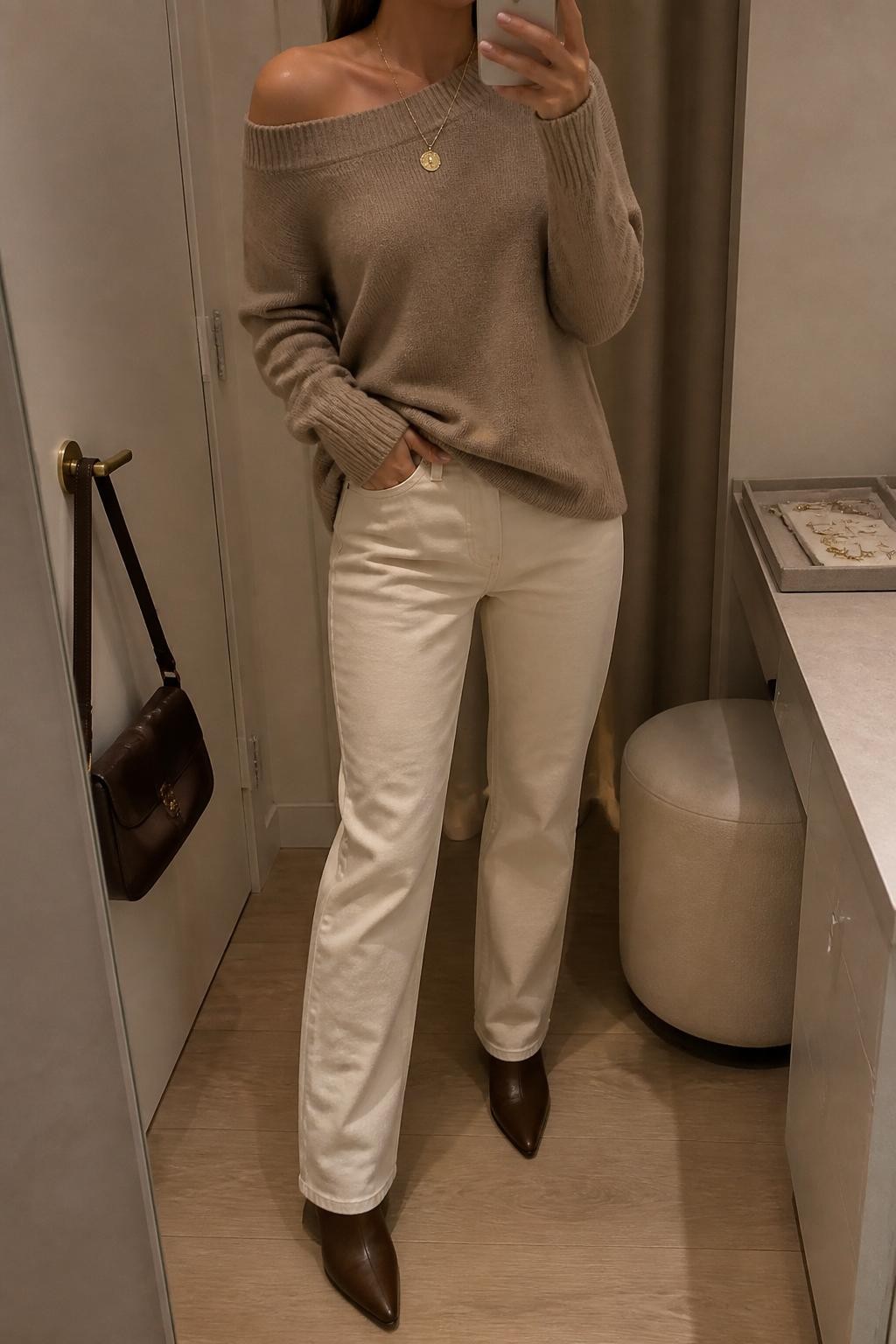 mirror selfie of a woman wearing a taupe lightweight off-shoulder sweater, ecru straight-leg jeans, brown pointed ankle boots, a gold pendant necklace, and a chocolate brown shoulder bag, clearly showing the same clothing items, colors, fabrics, and accessories listed in the Outfit Pieces section, holding a phone in front of a mirror, cropped at NECK so the face does not show, with a relaxed casual selfie pose, in a minimal dressing room, warm moody lighting, including a handbag hanging on a door handle and a jewelry tray on the vanity, latest iPhone photo quality.