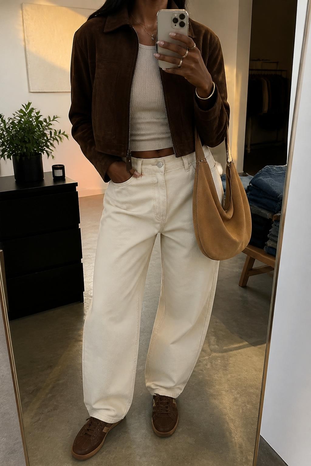 Mirror selfie of a Black woman with warm brown skin wearing a cropped chocolate brown suede jacket over a fitted oatmeal ribbed top, high-waisted ecru barrel-leg jeans, dark brown leather sneakers, and a soft tan suede hobo bag, clearly showing the exact clothing items, colors, textures, and accessories listed, holding a latest-model iPhone in front of a mirror, cropped at the neck so the face does not show, relaxed casual selfie pose, in a fashion studio mirror corner with evening golden-hour light, folded denim jeans on a bench and a small plant on a dresser, natural uncluttered setting, latest iPhone photo quality.