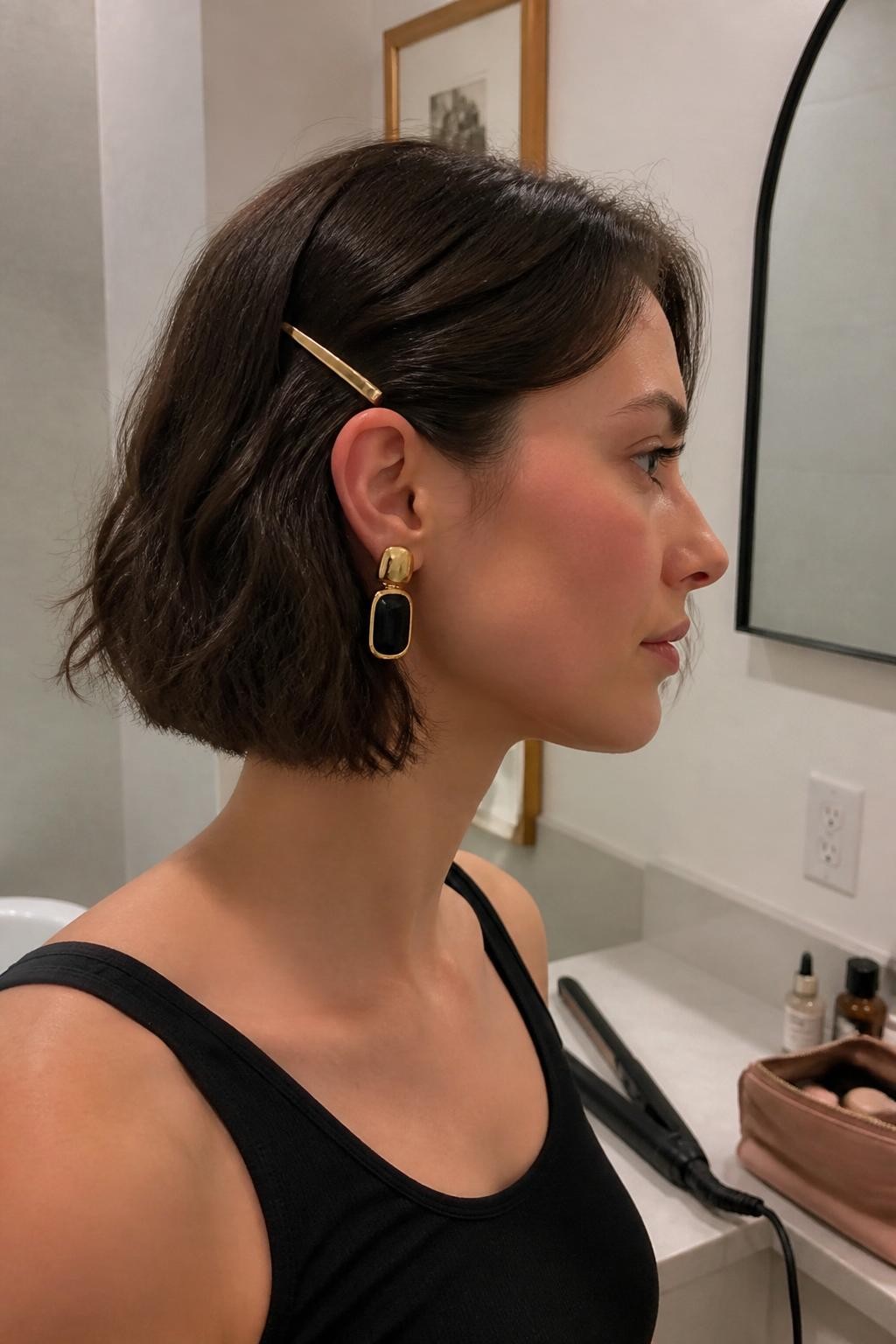 Image prompt: Photo of a woman wearing a chin-length tousled bob with loose flat-iron bends and one side pinned behind the ear with a slim gold barrette, side-profile view, bathroom vanity mirror setting, soft indoor lighting, flat iron on the counter and a makeup pouch nearby, statement earring visible on the pinned side, natural uneven texture, latest iPhone photo quality.