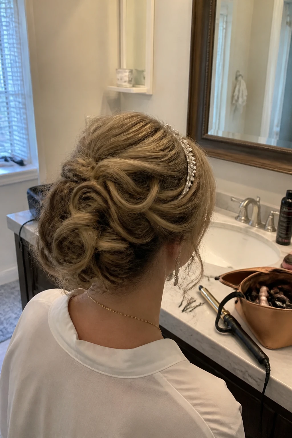 Image prompt

Photo of a woman wearing a soft curly French twist with gentle crown volume, loose tendrils around the face, and a delicate crystal-embellished headband, back-of-head view beside a bathroom vanity mirror, soft indoor lighting, containing a small lived-in detail such as long bobby pins near a makeup bag, a curling wand on the counter, and statement earrings peeking through, realistic getting-ready moment, latest iPhone photo quality.